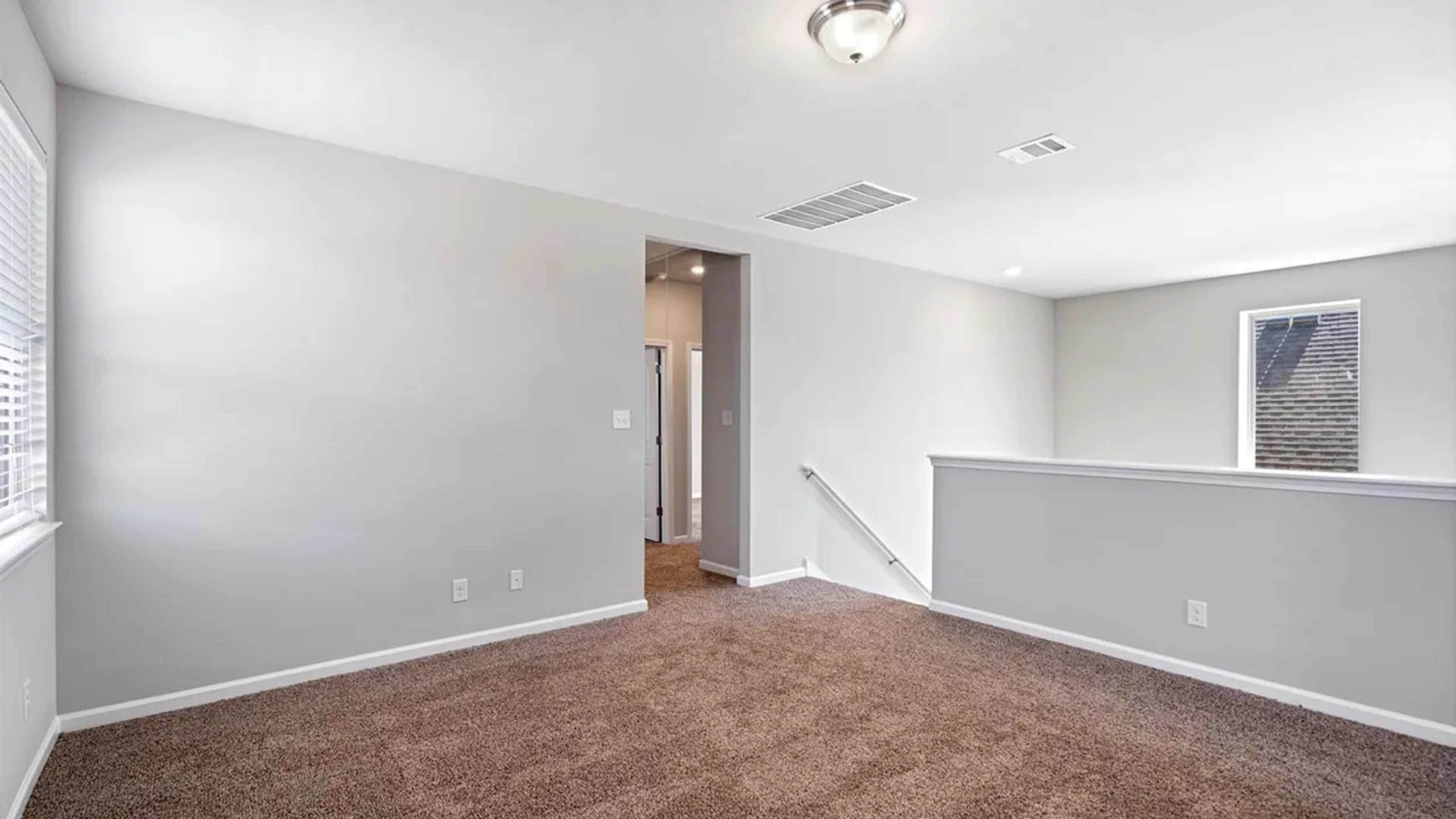 The image shows a spacious, empty room with beige carpet and light gray walls, featuring a ceiling light fixture and an opening leading to a hallway.