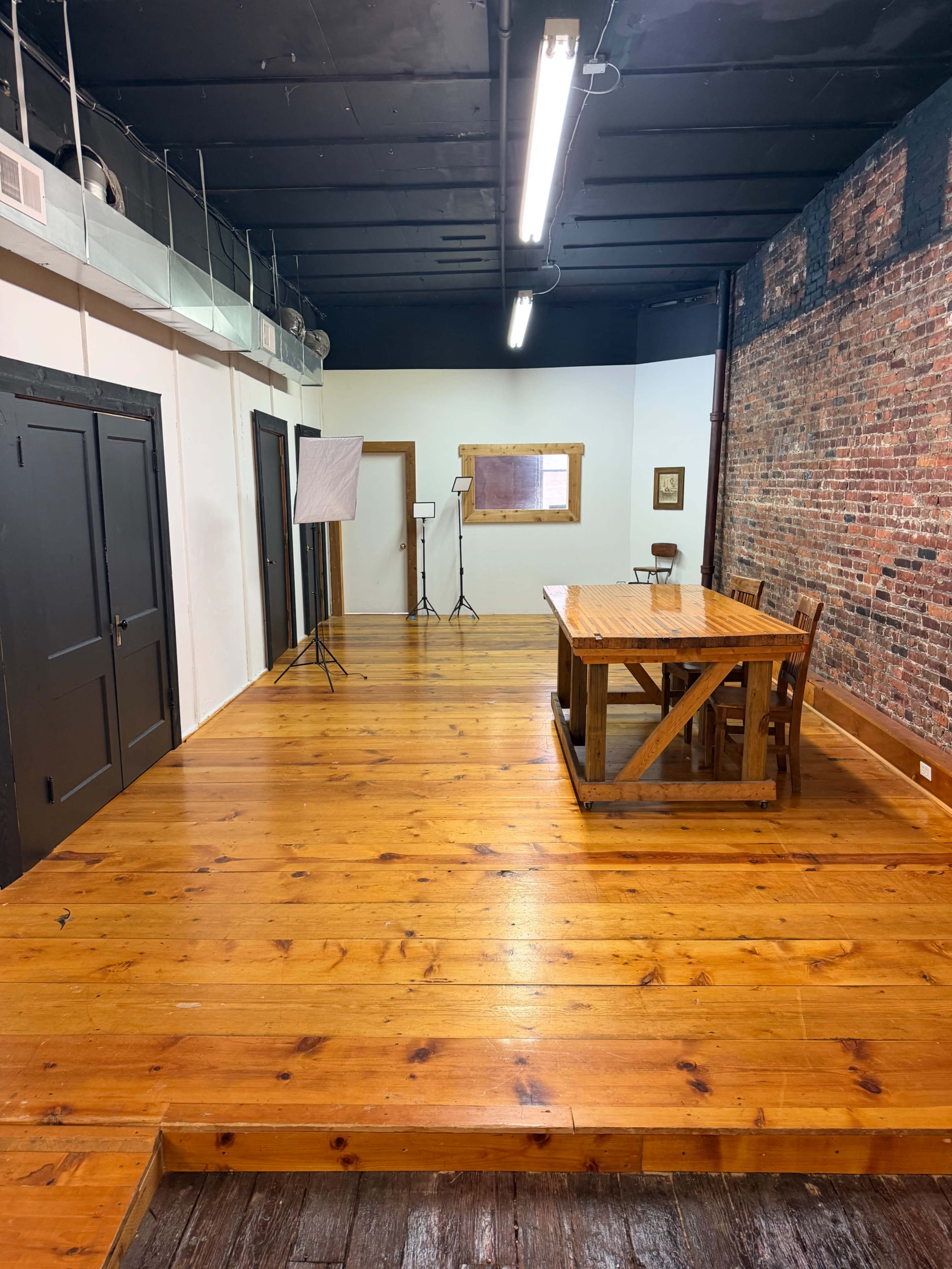 The image shows a spacious room with wooden floors, a large wooden table at the center, and exposed brick walls.