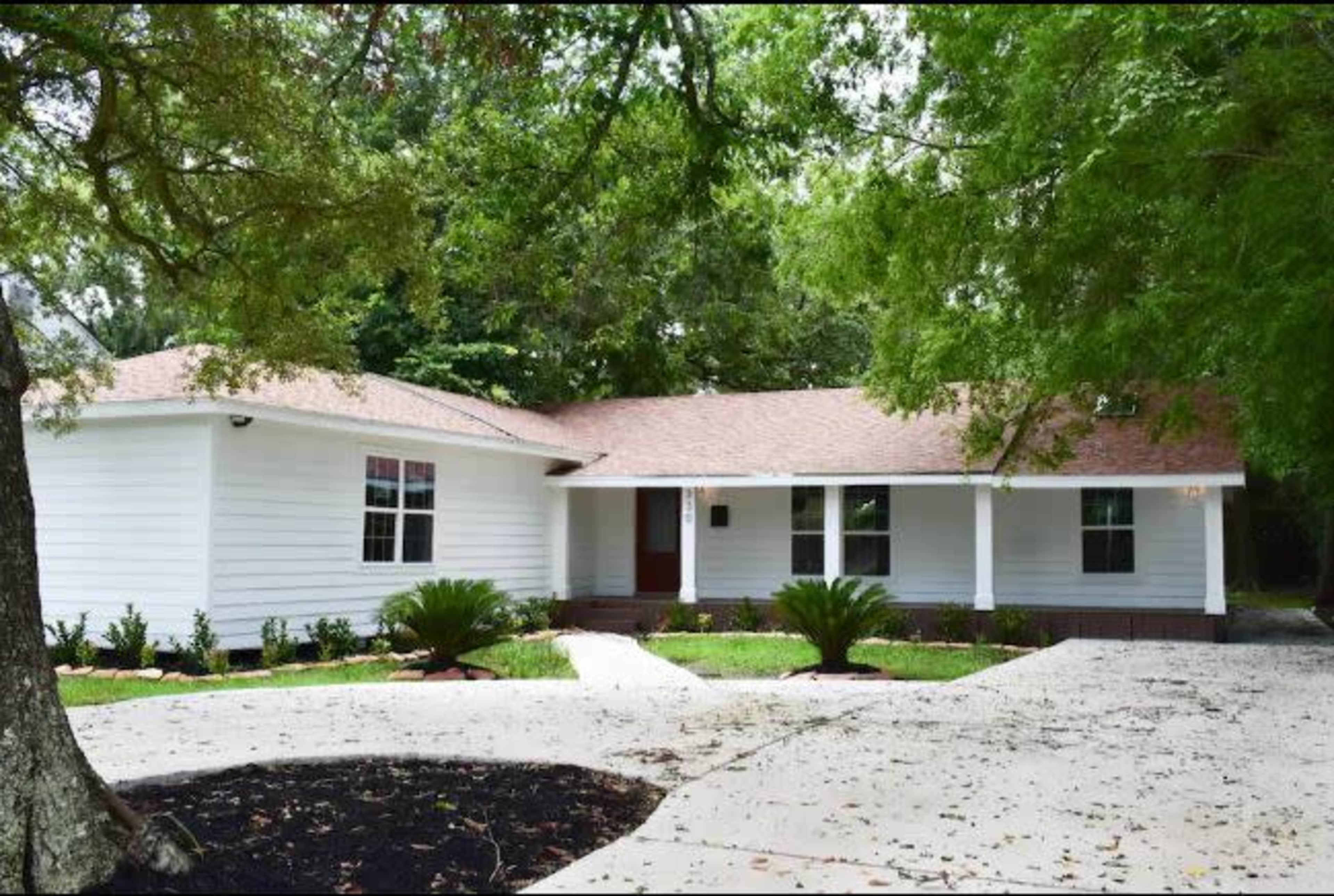 A single-story white house with a brown roof sits on a landscaped front yard featuring palm plants and a paved driveway.
