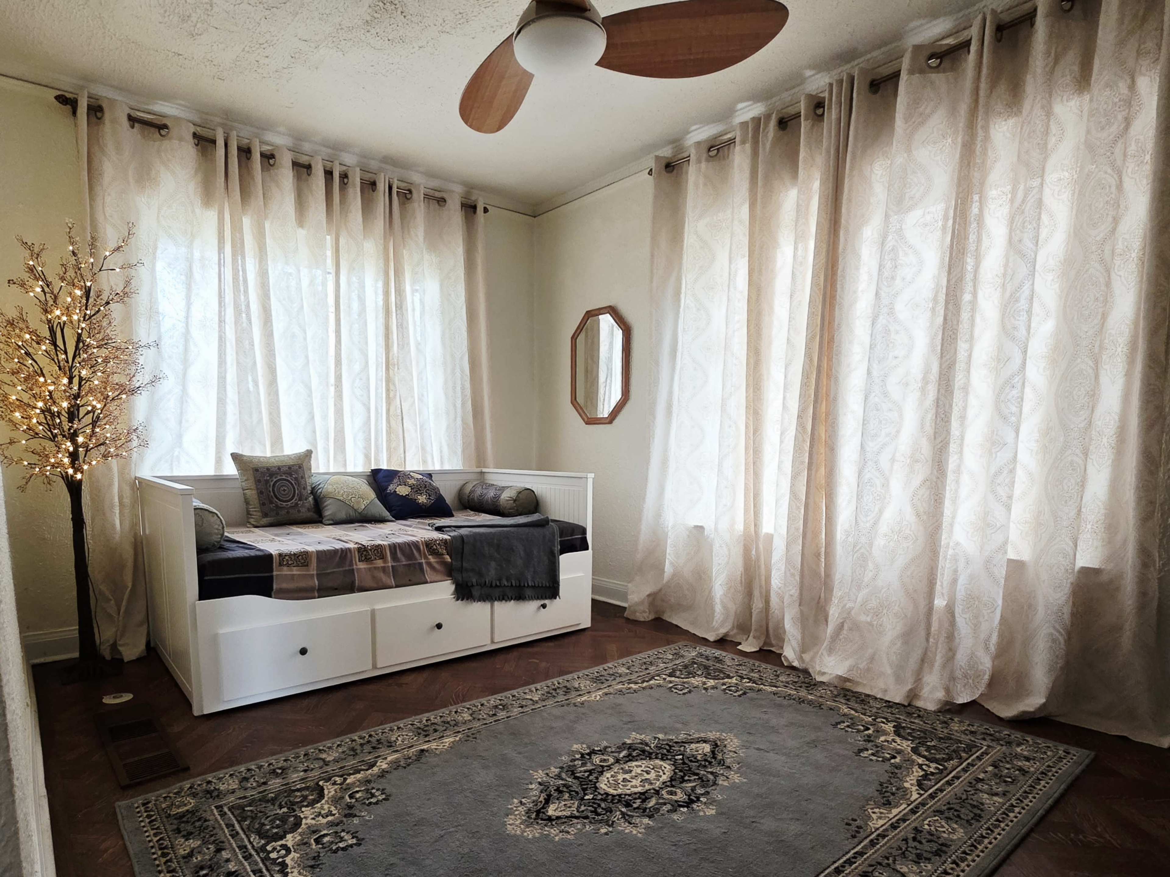The room features a daybed with decorative pillows, a large area rug, and sheer curtains allowing natural light to enter.