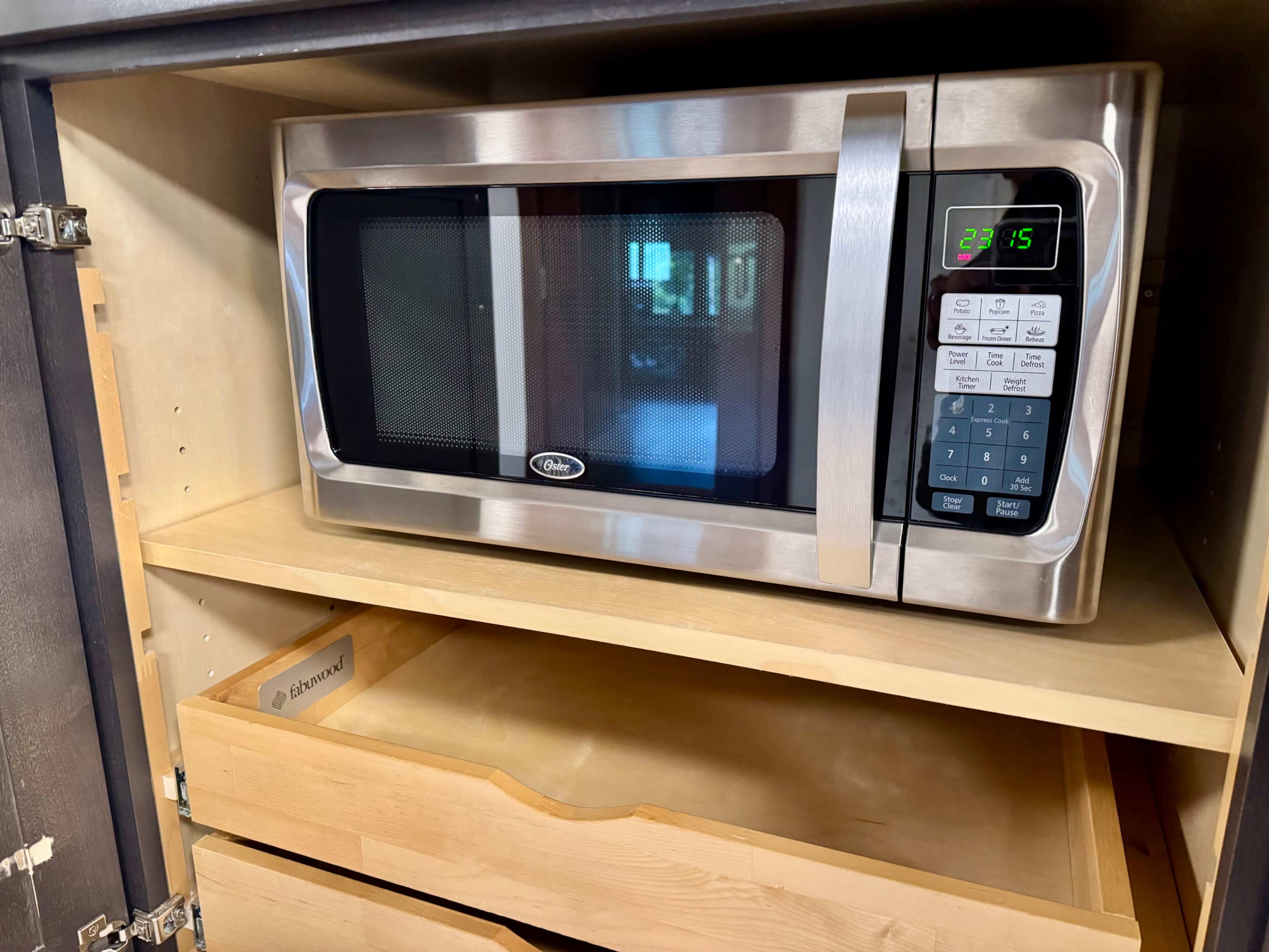 A stainless steel microwave is placed inside a wooden kitchen cabinet with a drawer below it.