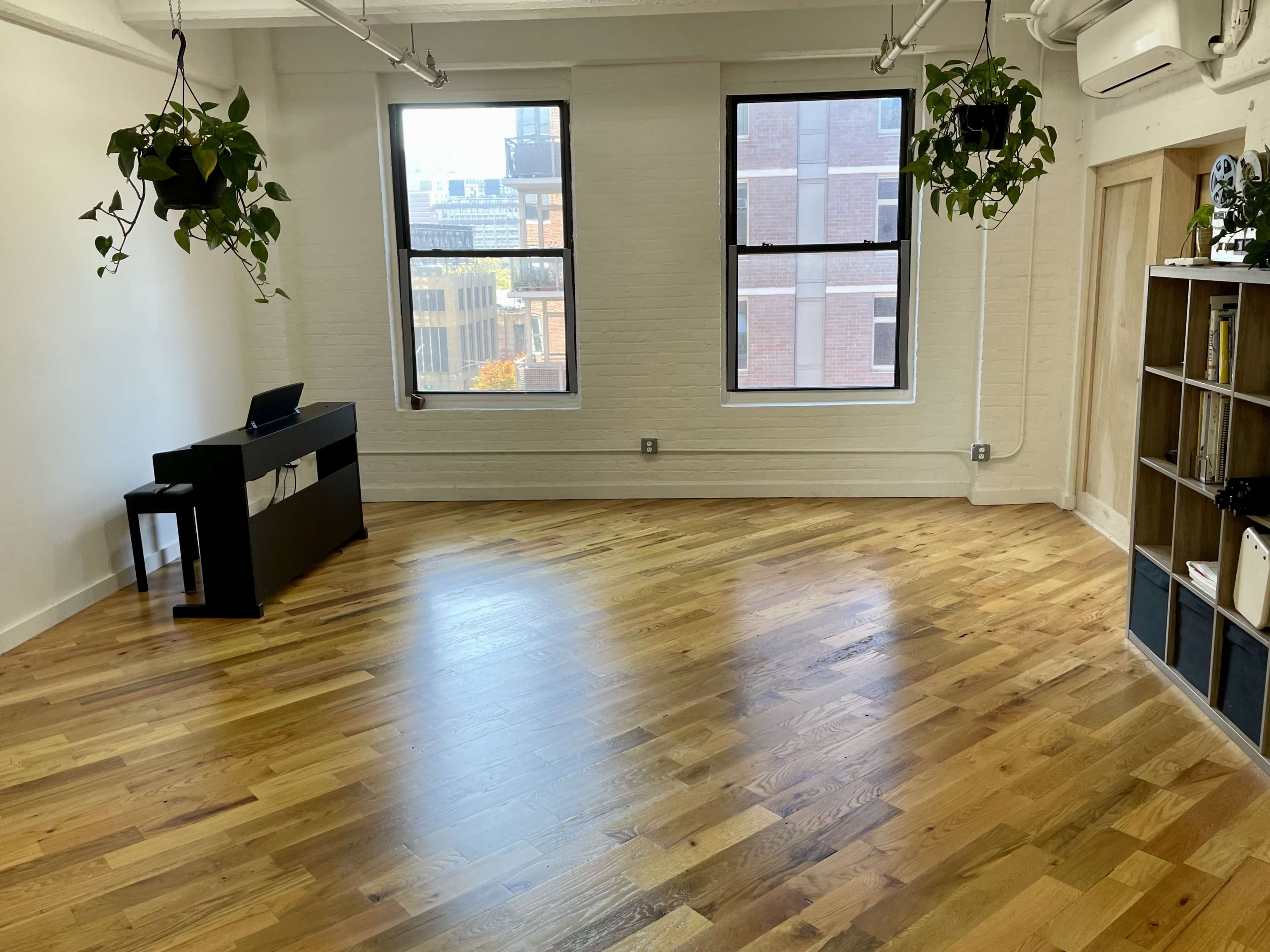 A spacious, well-lit room with hardwood flooring, a black furniture piece against one wall, and plants hanging near the windows.