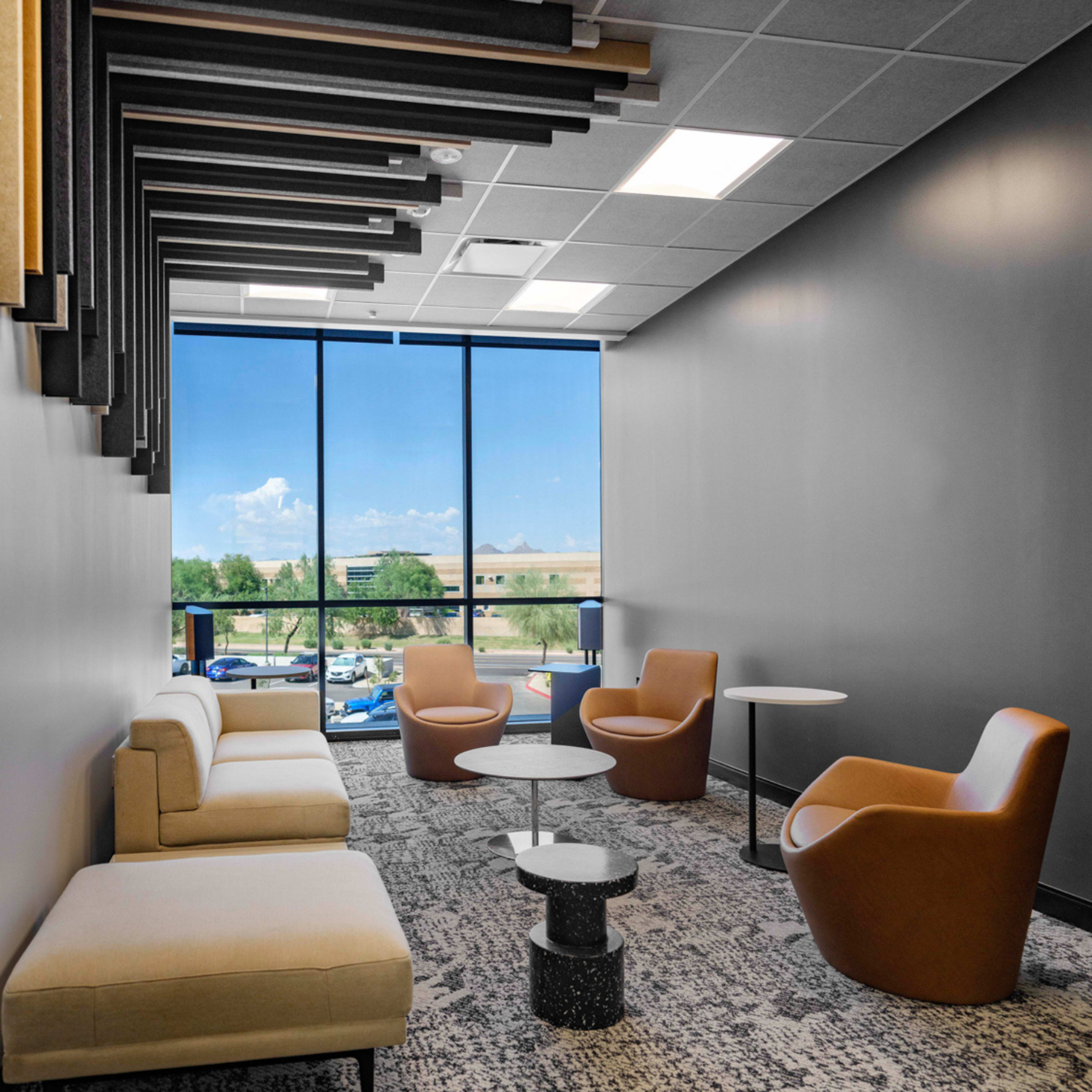 The image shows a modern lounge area with a sofa, two armchairs, and small tables, featuring large windows that provide a view of the outdoors.