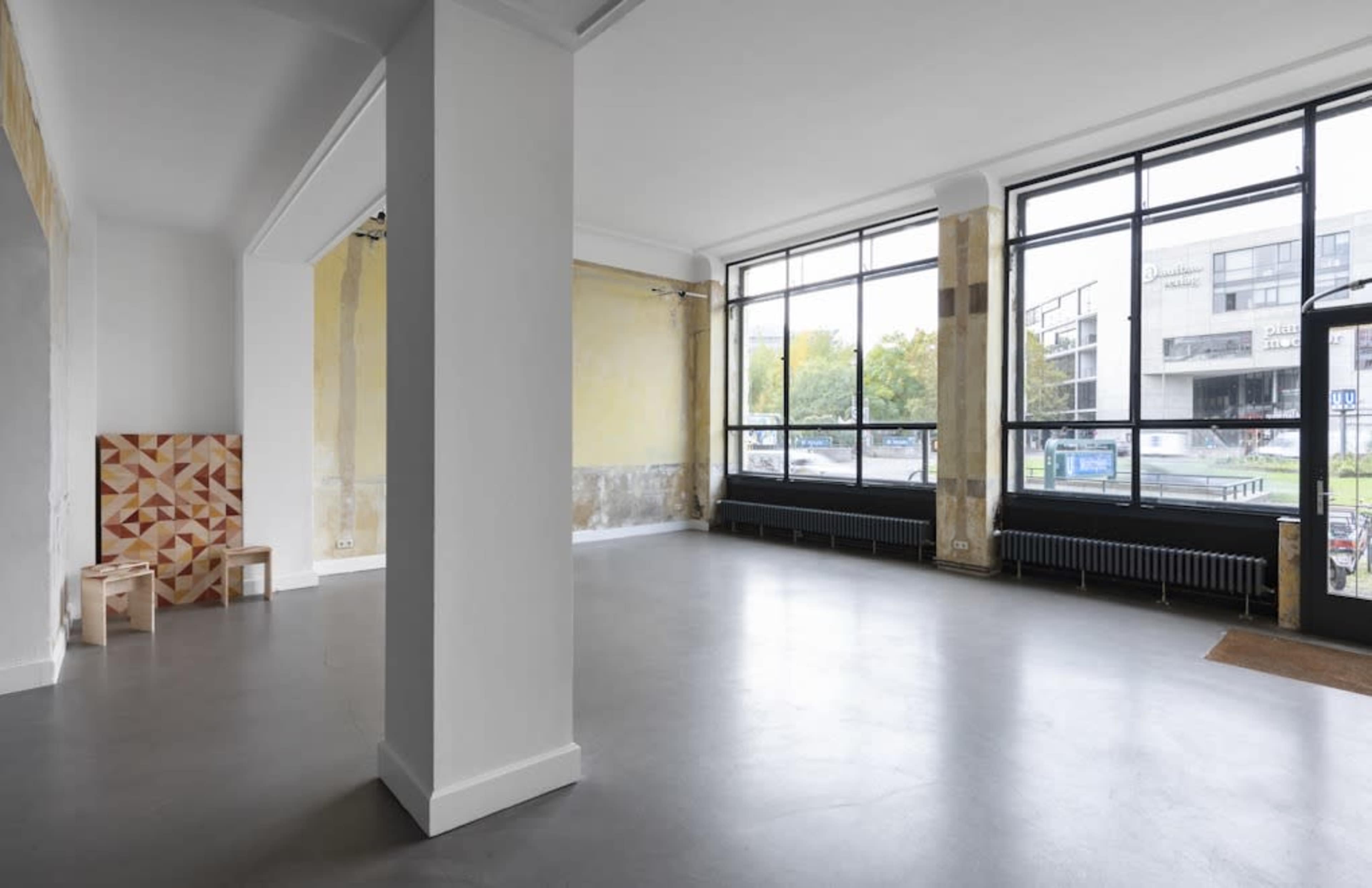 An empty interior space with large windows, exposed walls, and minimal furnishings, featuring a geometric wooden chair set against one wall.