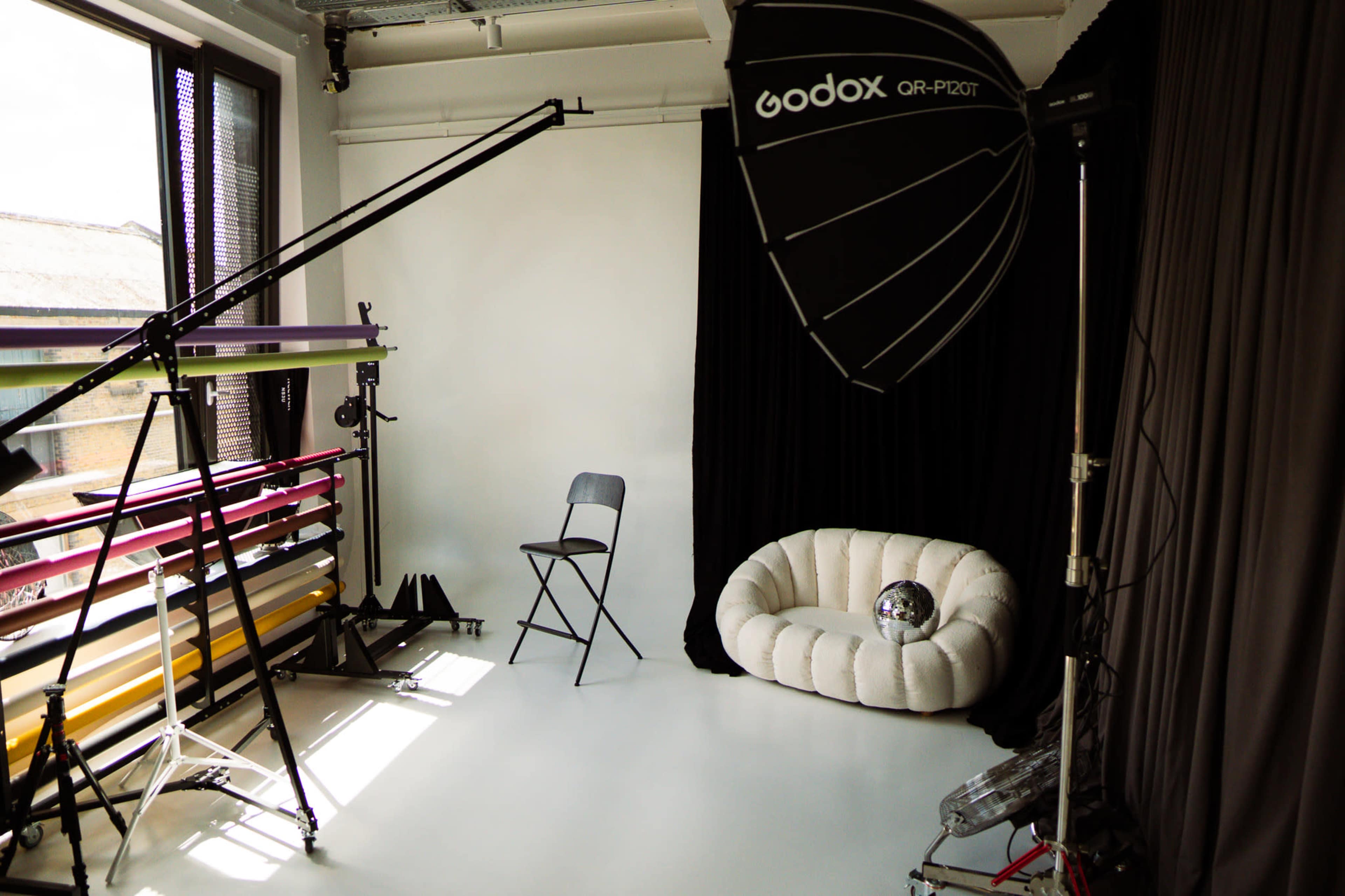 The image shows a photography studio with a light setup, a folding chair, and a white couch against a backdrop.