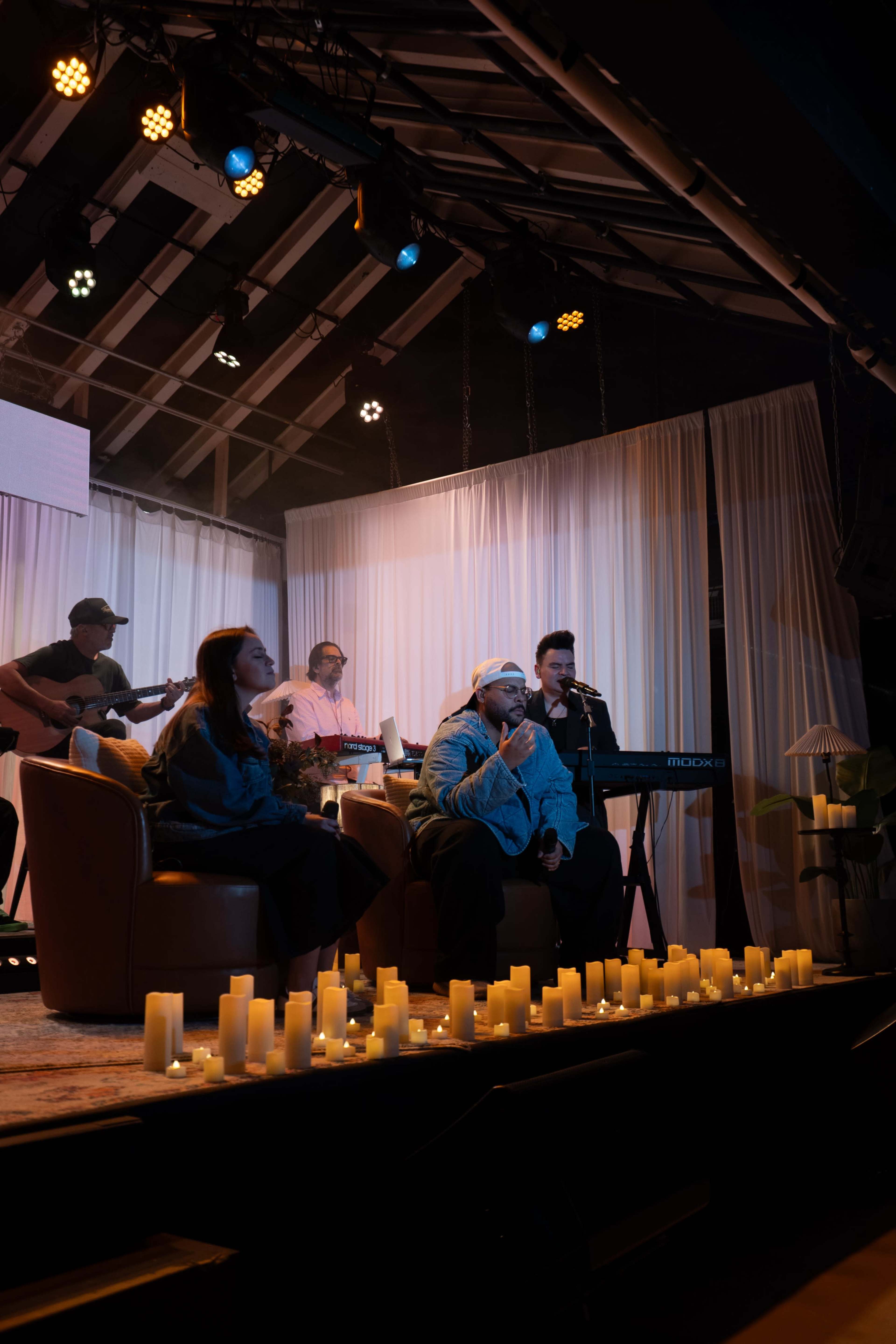 A group of musicians performs on a stage adorned with candles, surrounded by soft drapery and dim lighting.