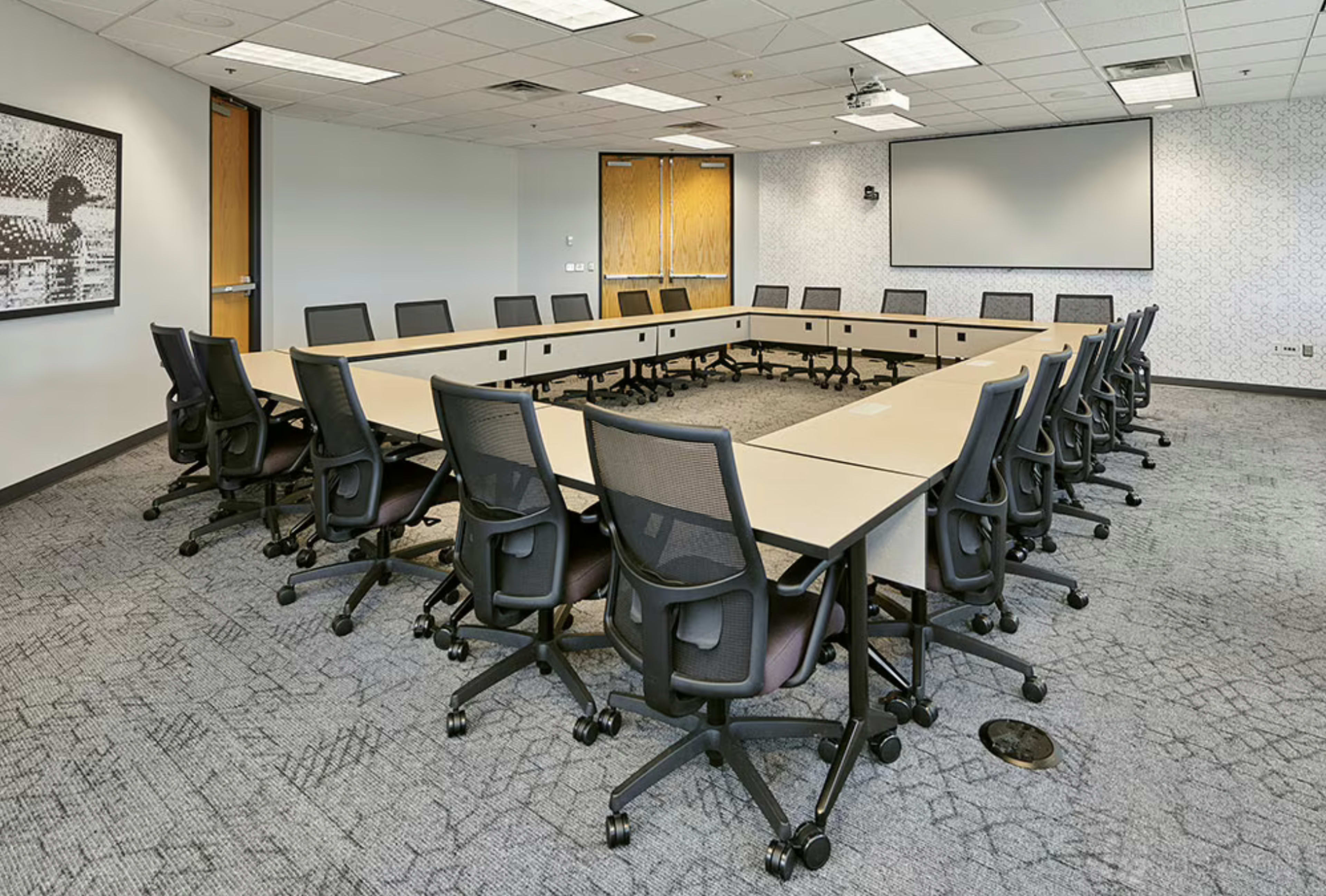 A large conference room features a U-shaped arrangement of tables surrounded by black office chairs and equipped with a projector screen at the front.