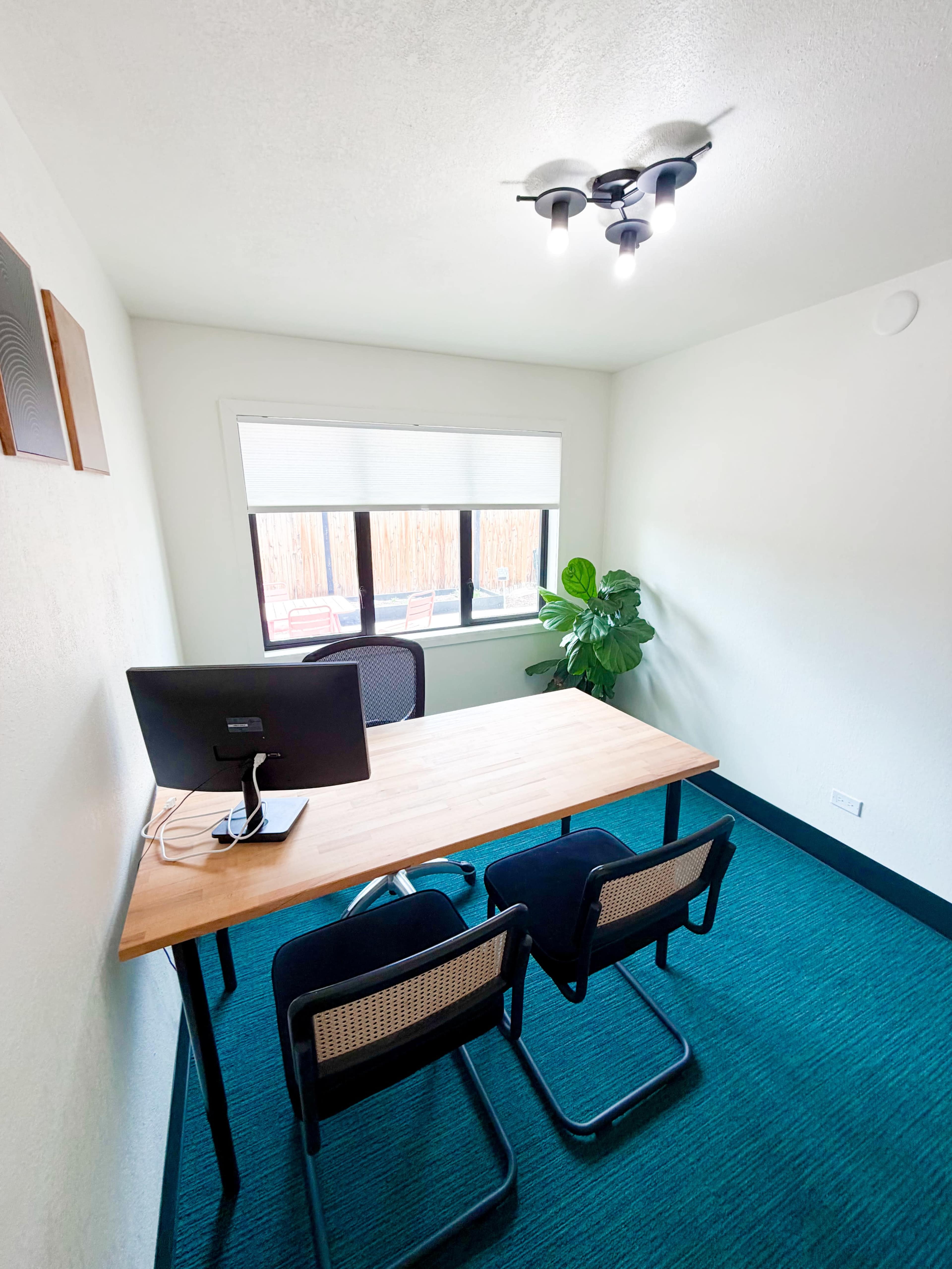 The image shows a small office space with a wooden desk, two chairs, a computer monitor, and a large window allowing natural light, alongside a potted plant in the corner.