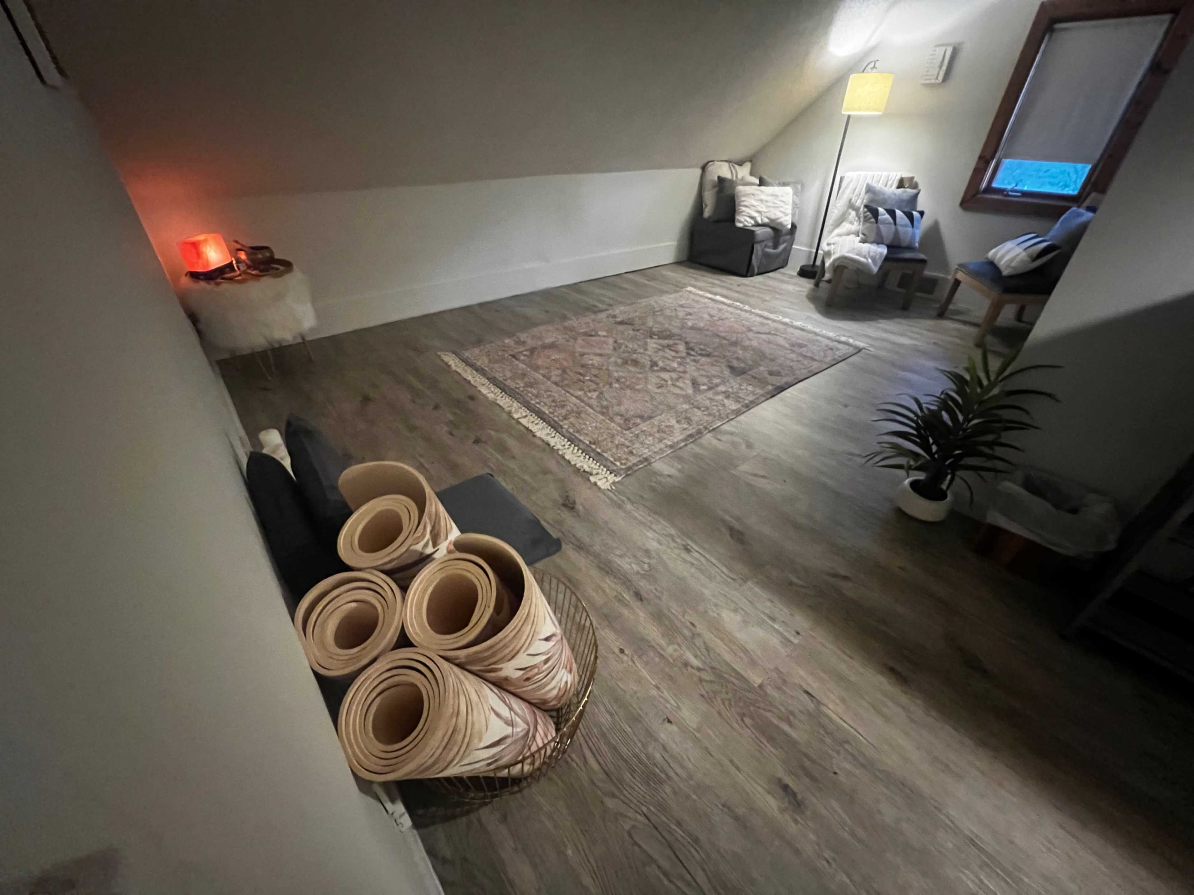 A spacious, well-lit attic room features a woven rug, a small sofa, a chair, and neatly rolled mats in a basket.
