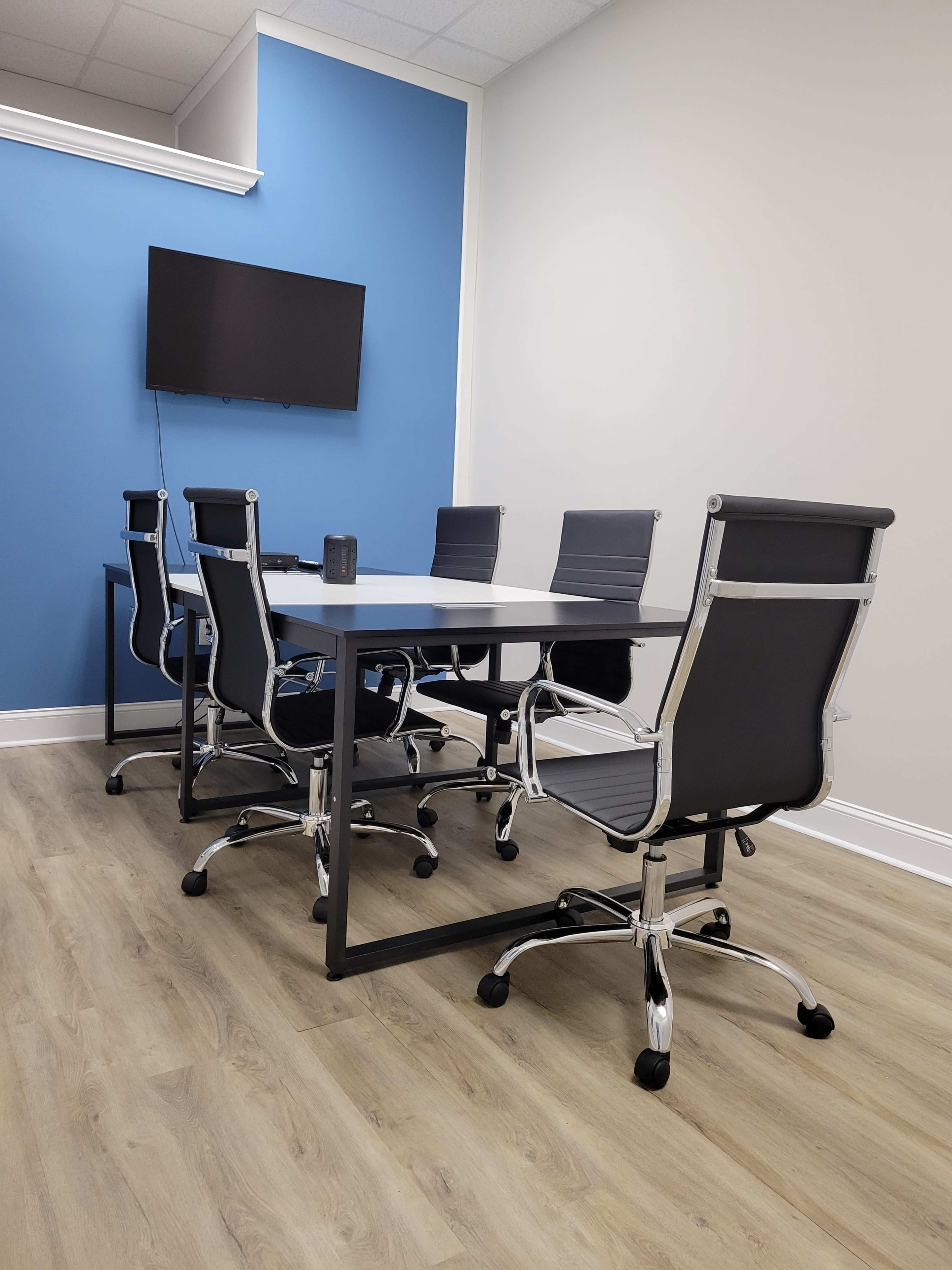 A modern conference room features a rectangular table surrounded by six rolling chairs and a wall-mounted television.