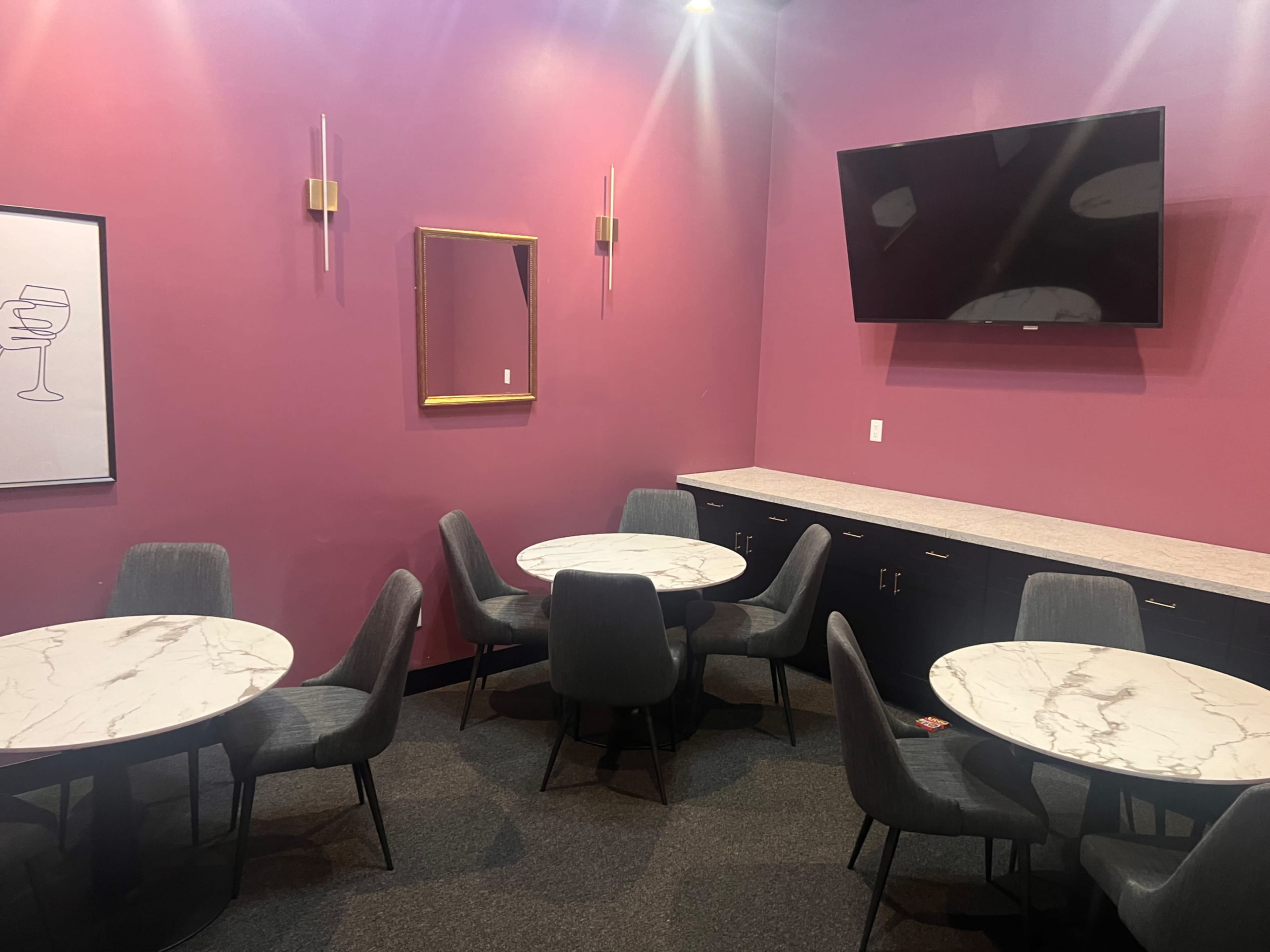 The image shows a seating area with several round marble tables and gray chairs against a purple wall, featuring a flat-screen TV and a mirror.