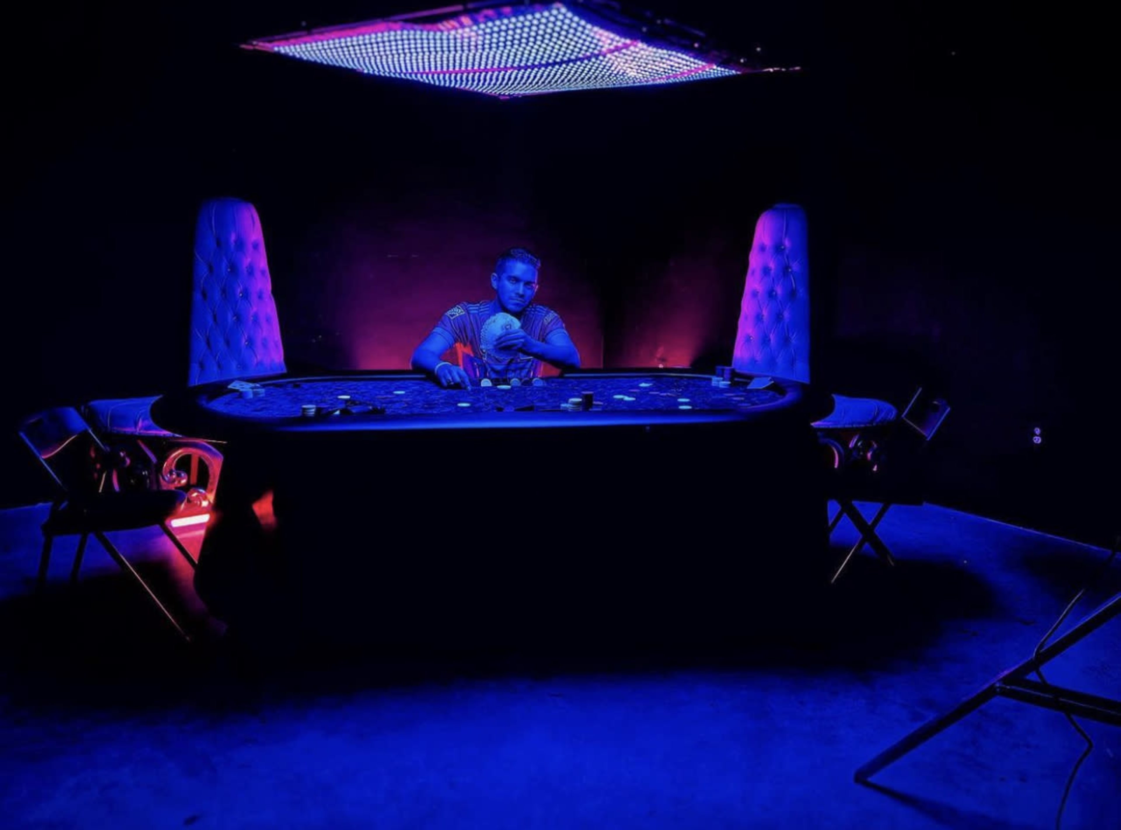 A lone player sits at a poker table under a brightly lit ceiling in a dimly lit room.