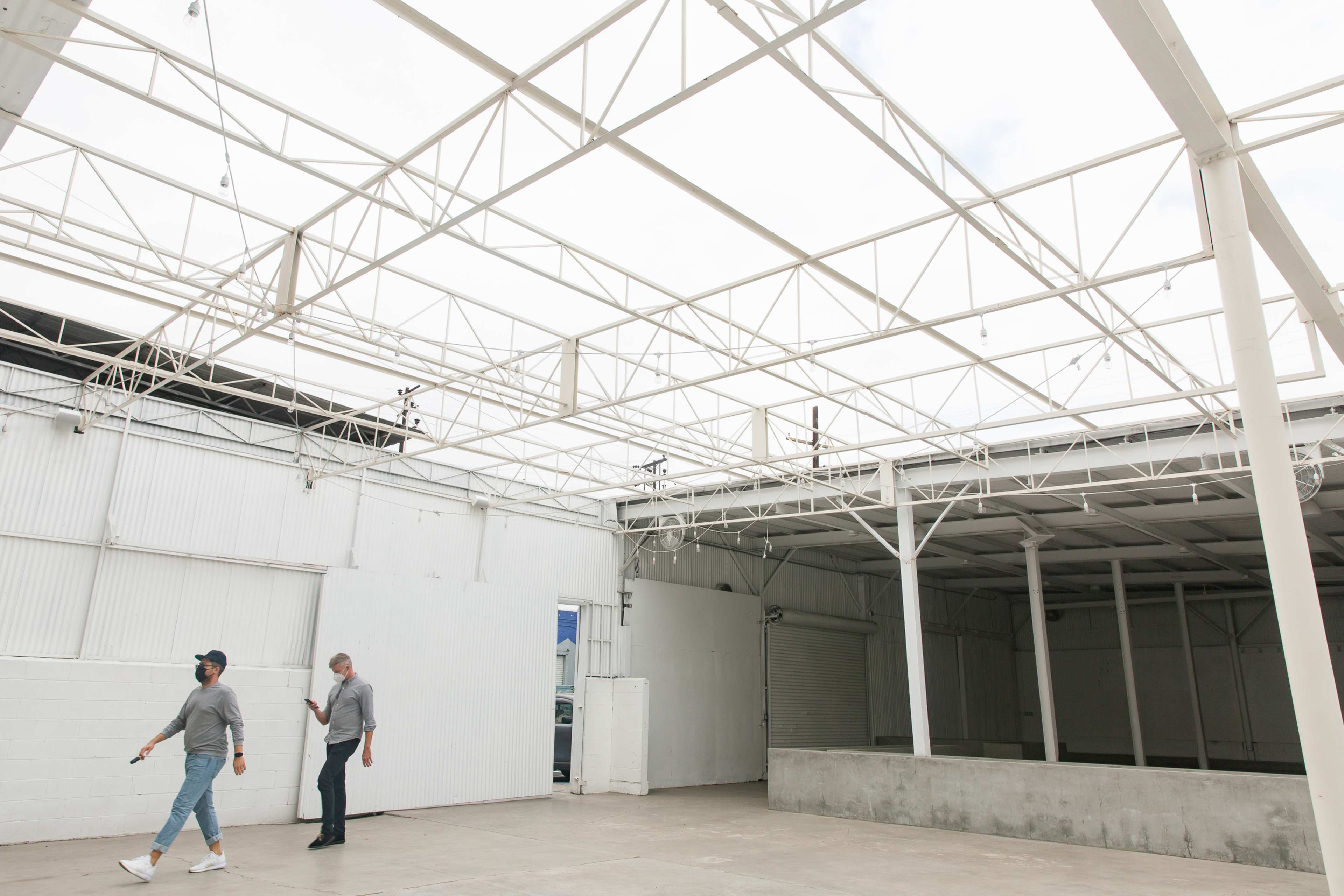 Two men walk through a spacious, unfinished industrial area with a high, open ceiling and white walls.