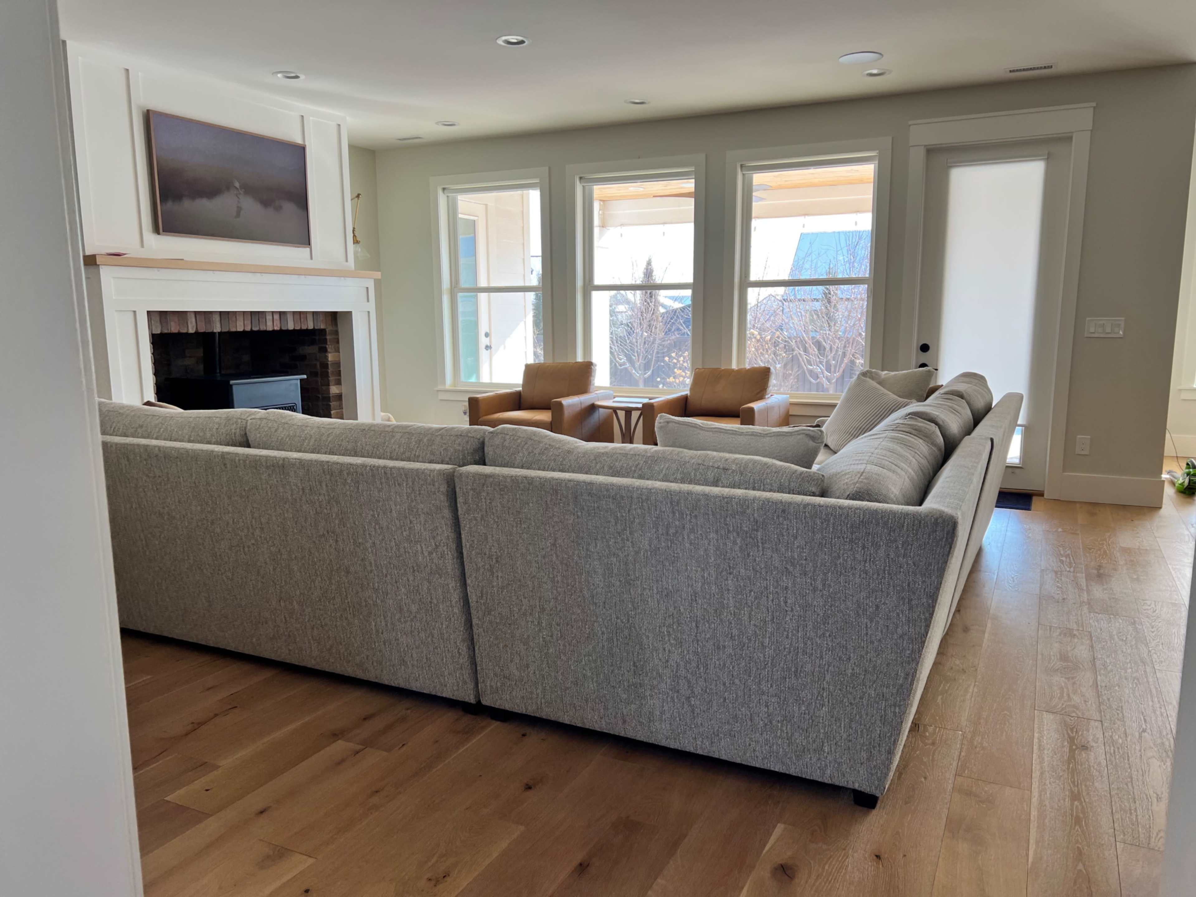 The living room features a large, L-shaped gray sofa facing a fireplace, with two armchairs and large windows providing natural light.