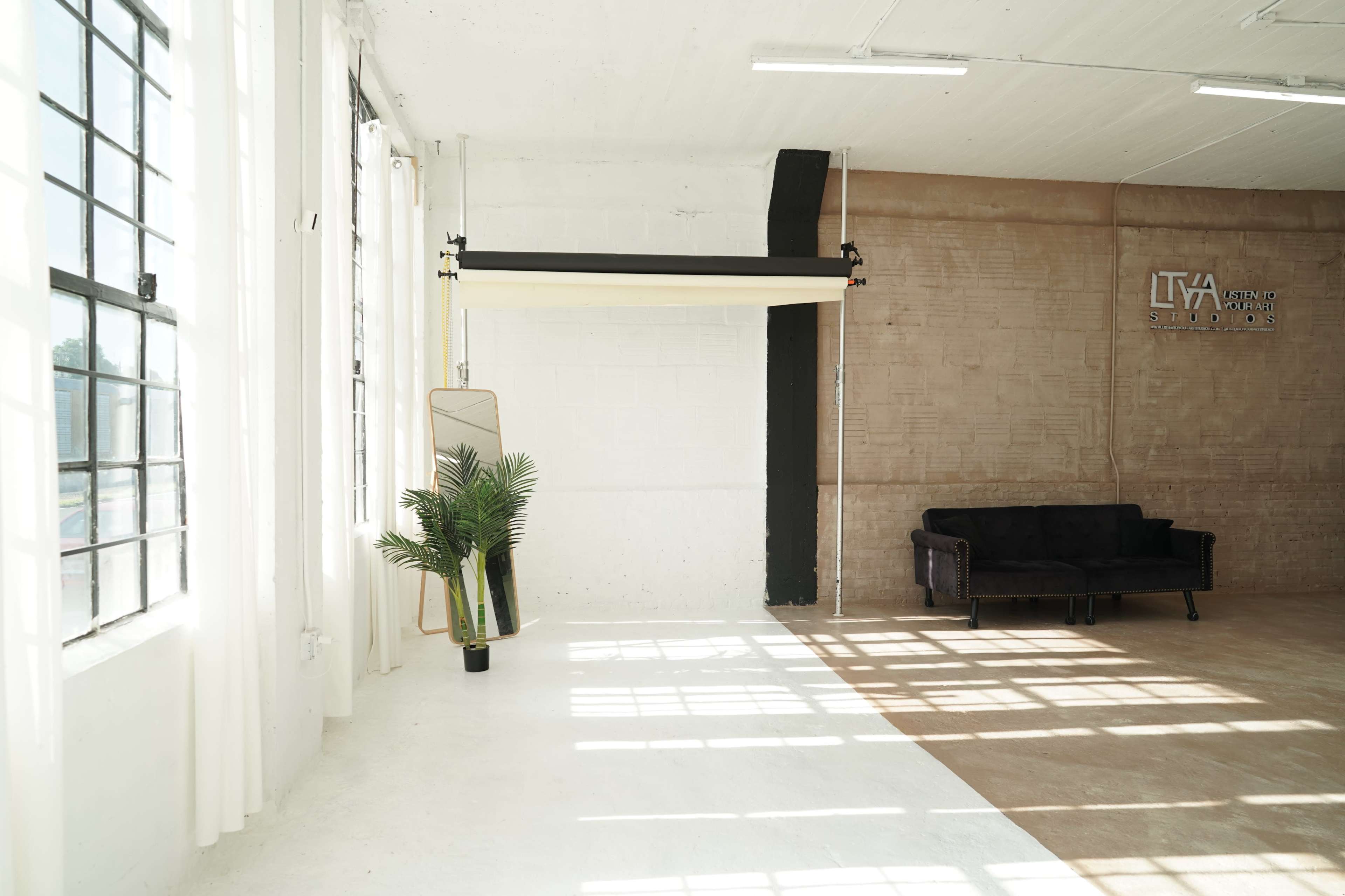 The image shows a bright studio space with large windows, a black couch, a potted plant, and a backdrop support system.