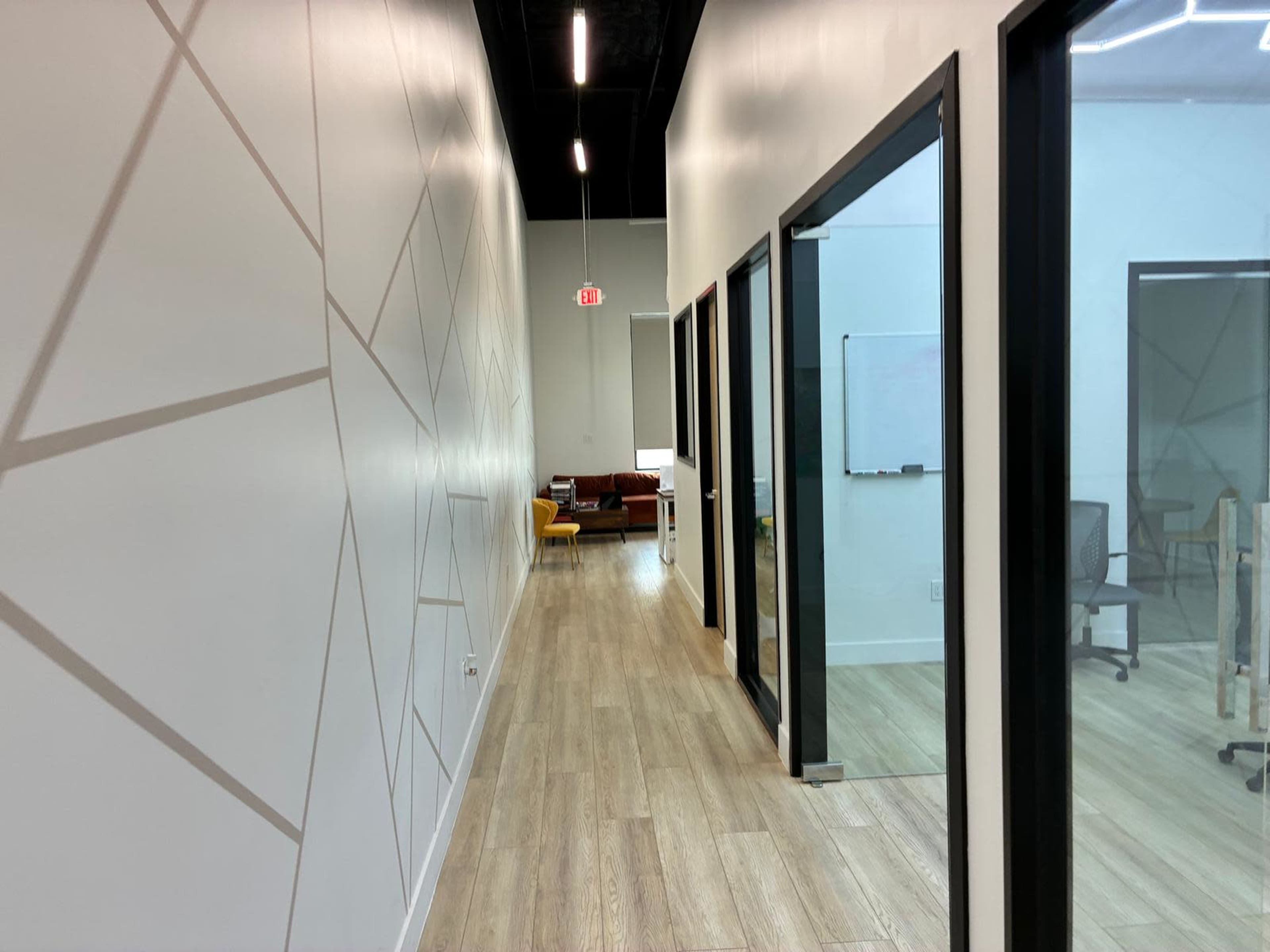 The image shows a modern hallway with light wood flooring, featuring glass-walled office spaces on either side and a seating area at the end.