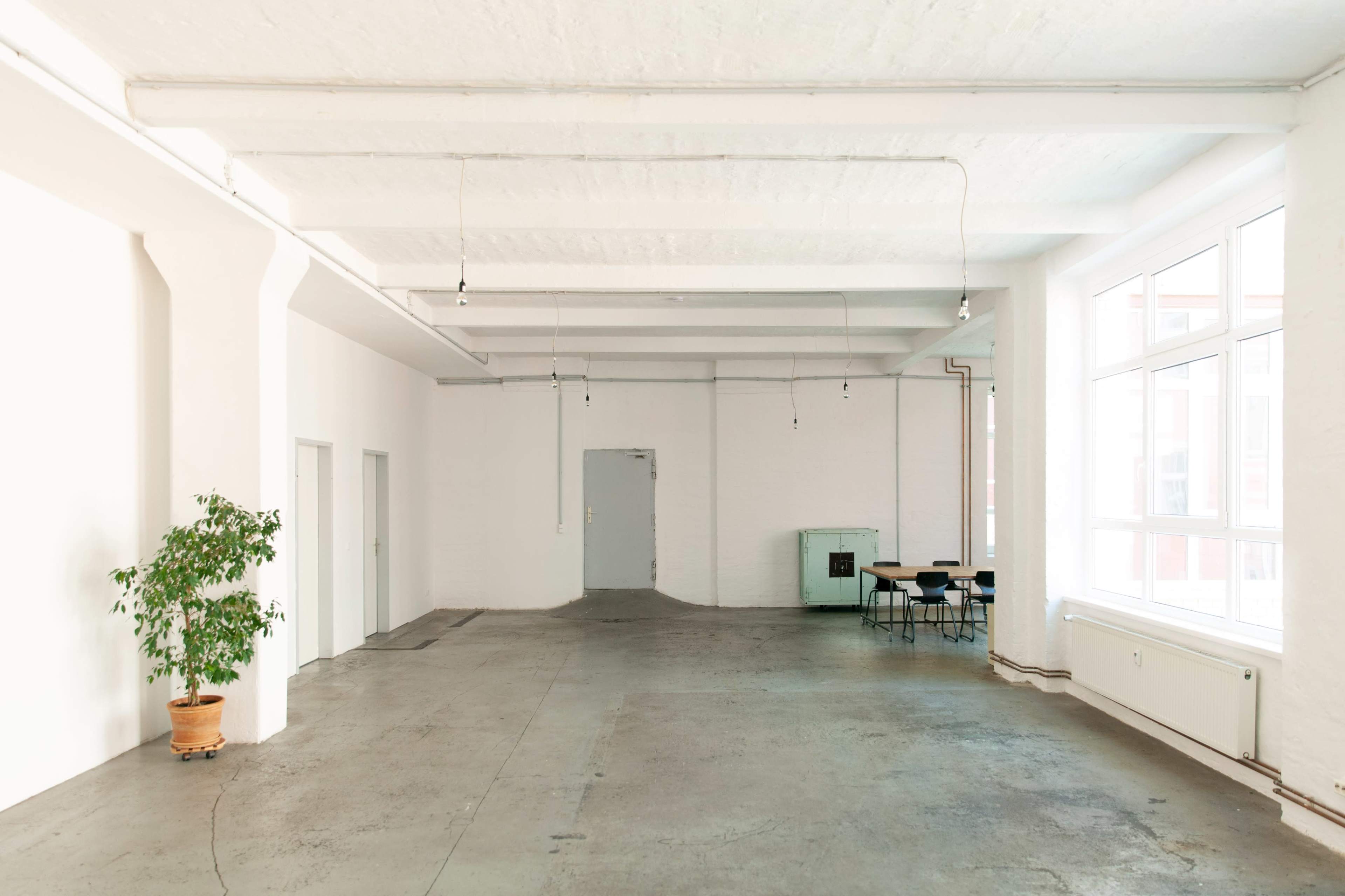 The image shows a spacious, well-lit room with concrete flooring, white walls, and minimal furnishings, including a small table and chairs, and a potted plant.