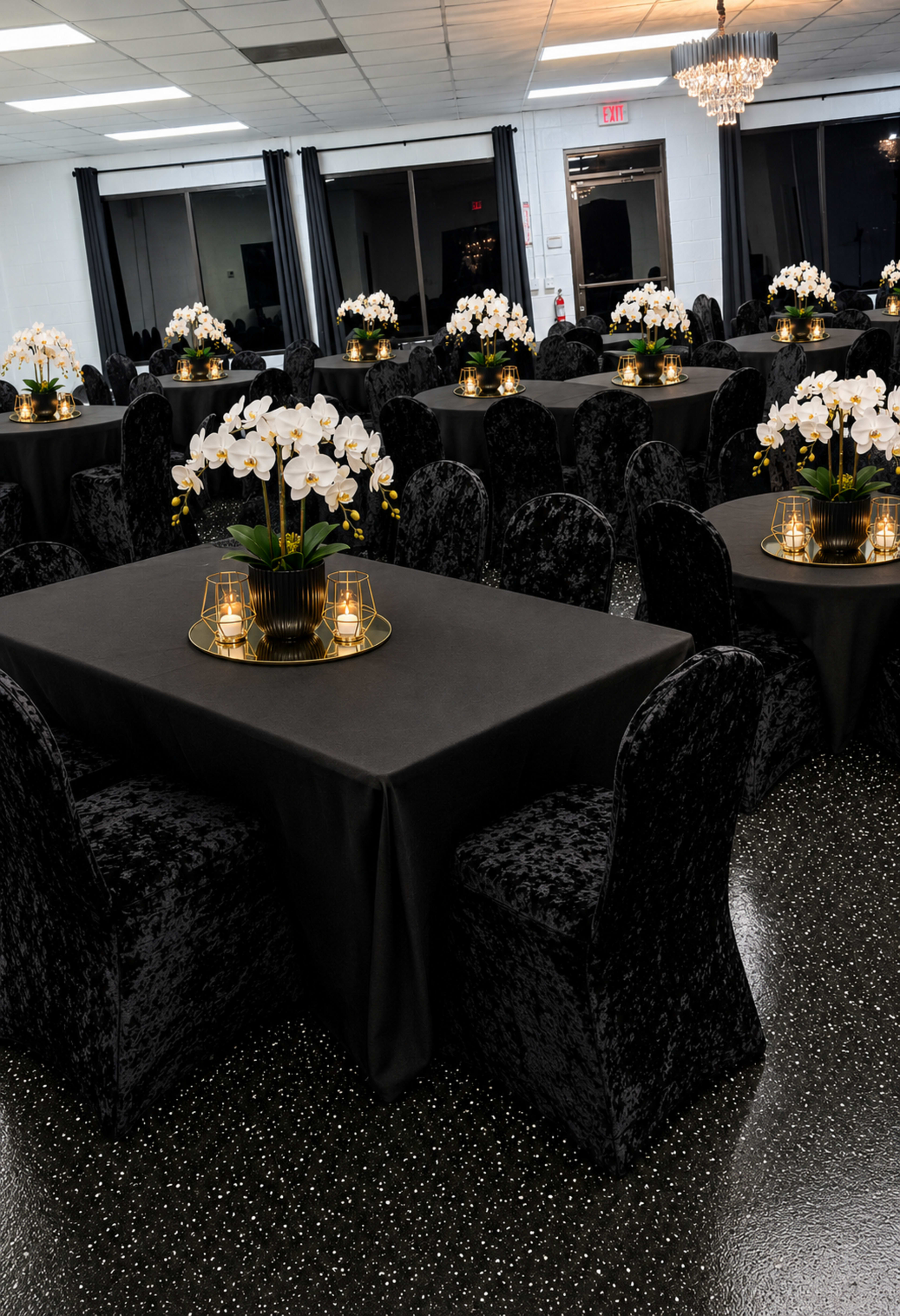 A banquet hall features several tables covered in black cloth, adorned with floral centerpieces and illuminated by decorative lanterns.