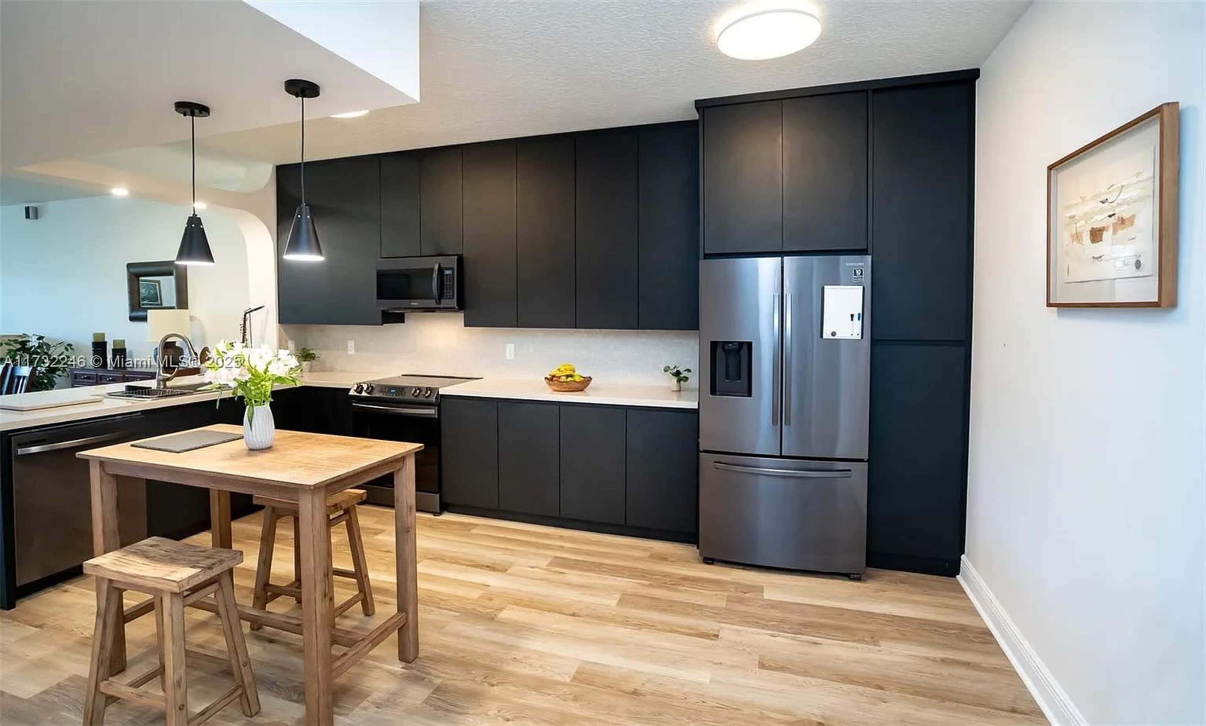 The image shows a modern kitchen featuring dark cabinetry, stainless steel appliances, and a wooden dining table with stools.