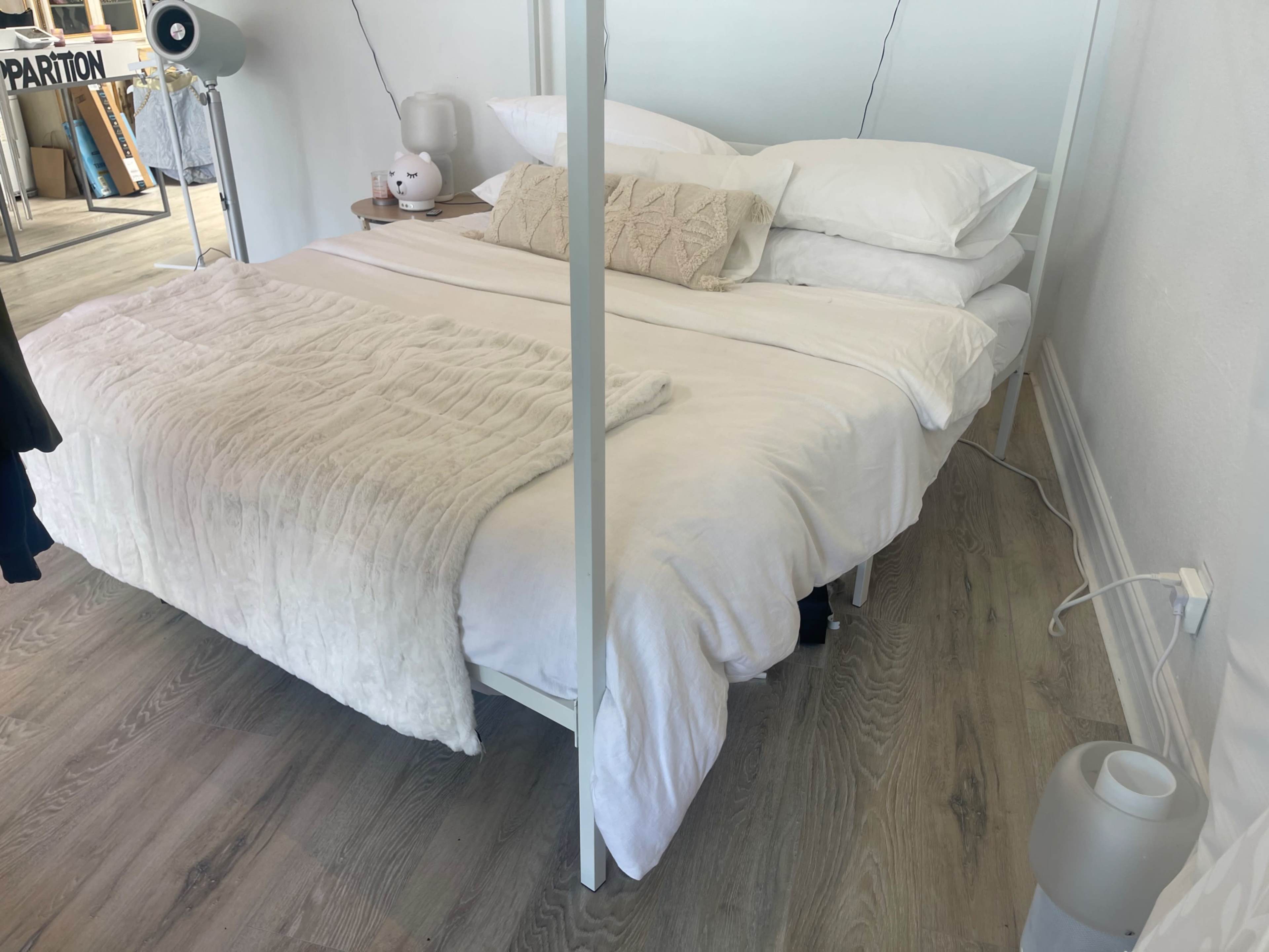 A white bed with layered bedding and pillows is positioned in a minimalist bedroom with wooden flooring.
