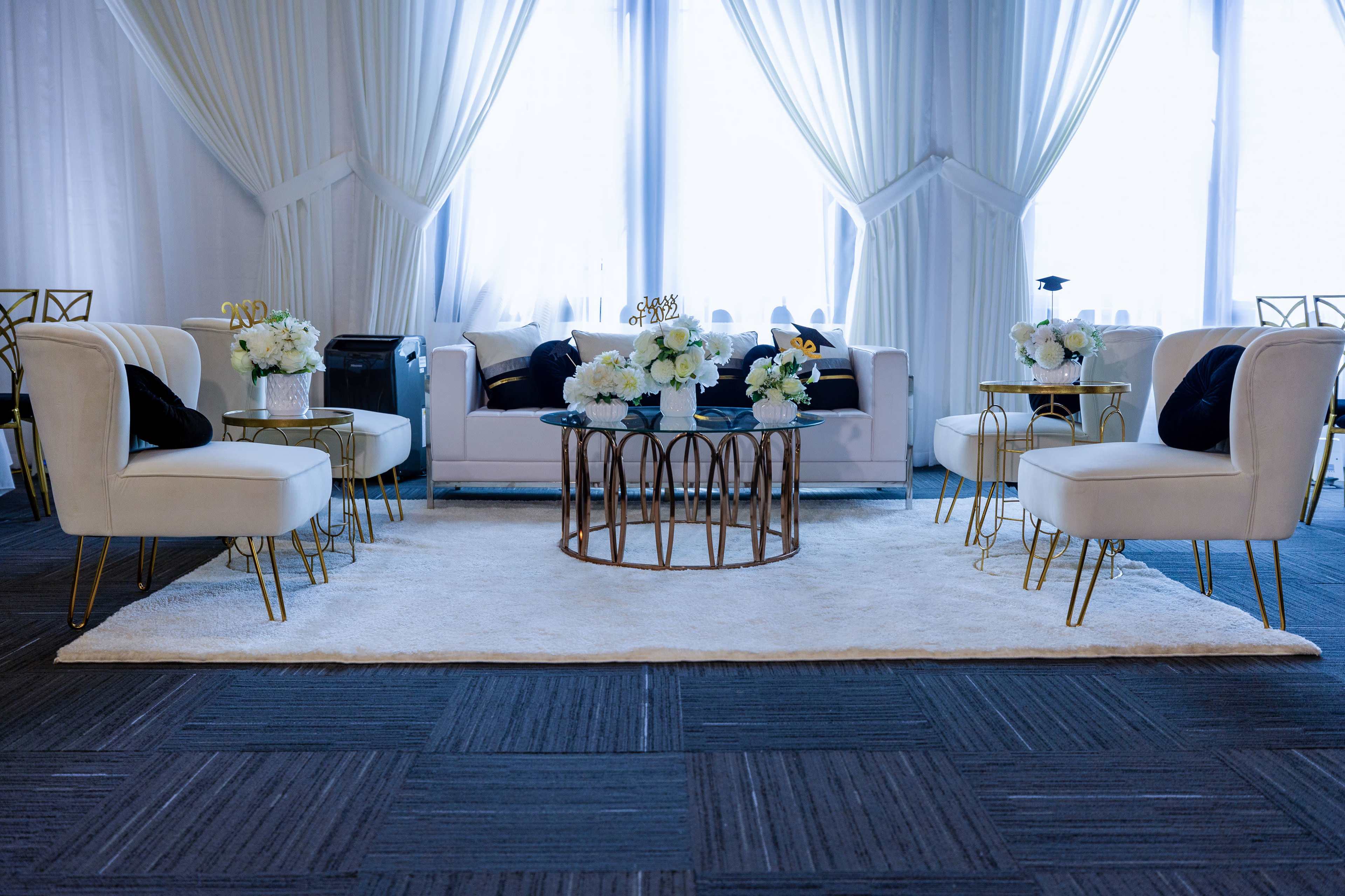 A stylish lounge area with white seating, a round coffee table, floral arrangements, and large windows covered by sheer curtains.