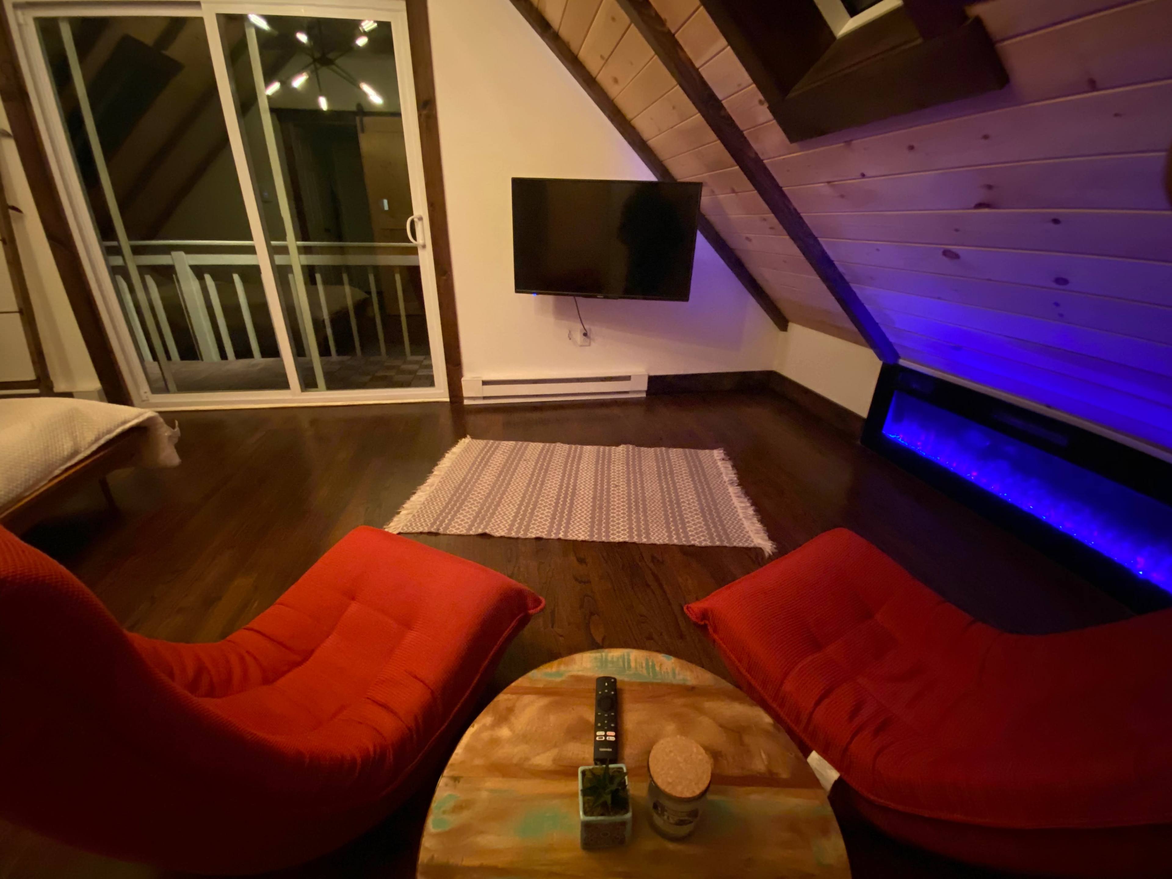 A cozy, triangular room with wood-paneled walls, featuring two red lounge chairs facing a wooden coffee table, a television mounted on the wall, and a lighted area near the floor.