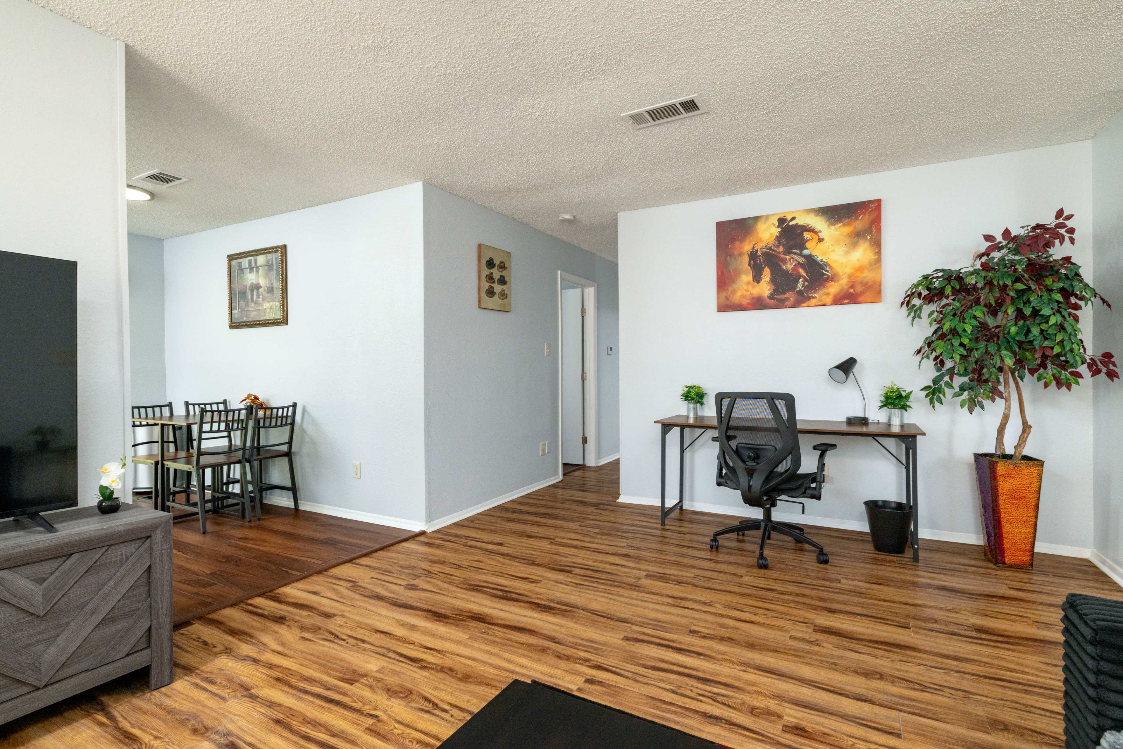 The image shows a room with wooden flooring, a desk with a chair, a television, a dining area, and a decorative plant.