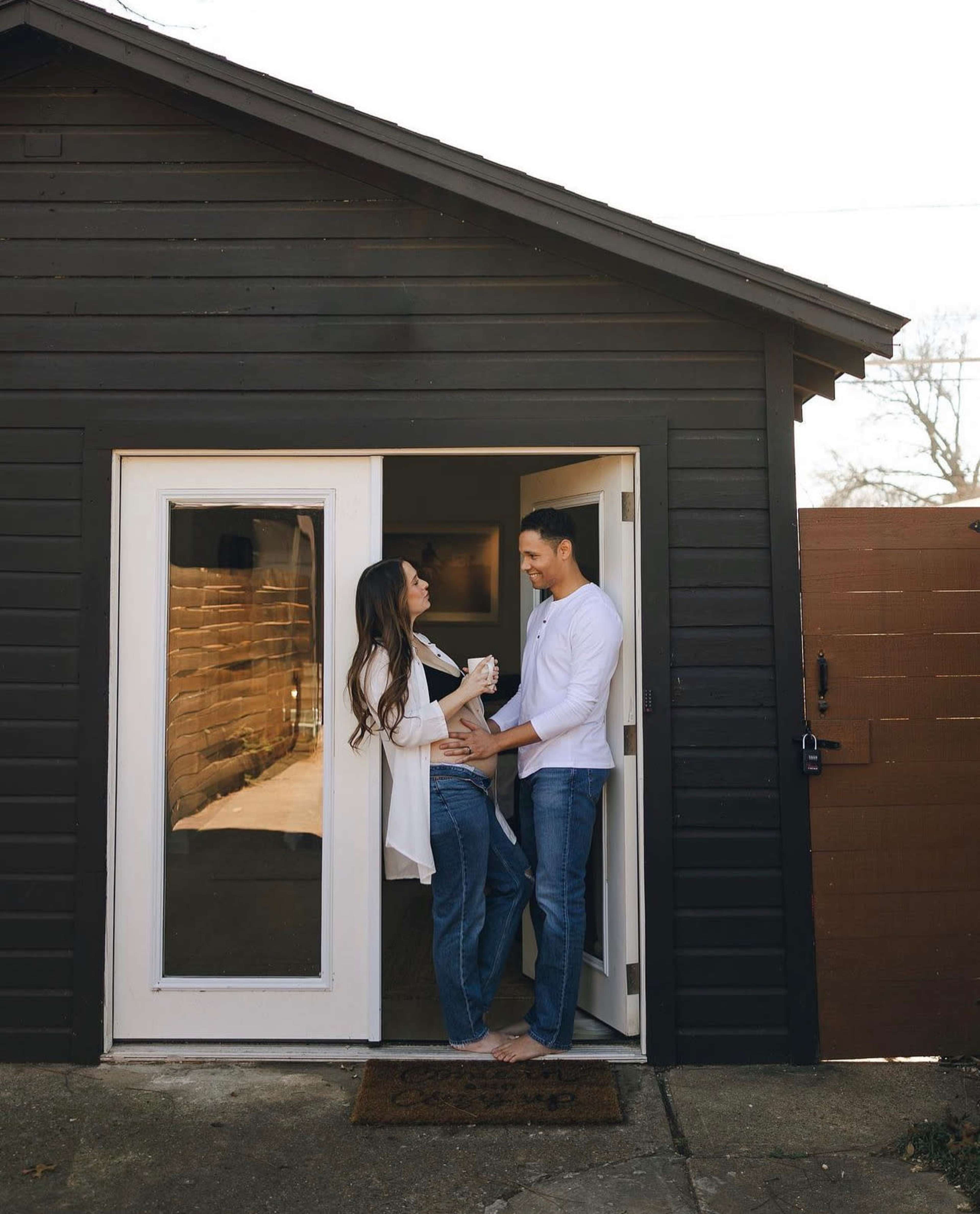 A couple stands playfully in a doorway of a black wooden house, enjoying each other's company.
