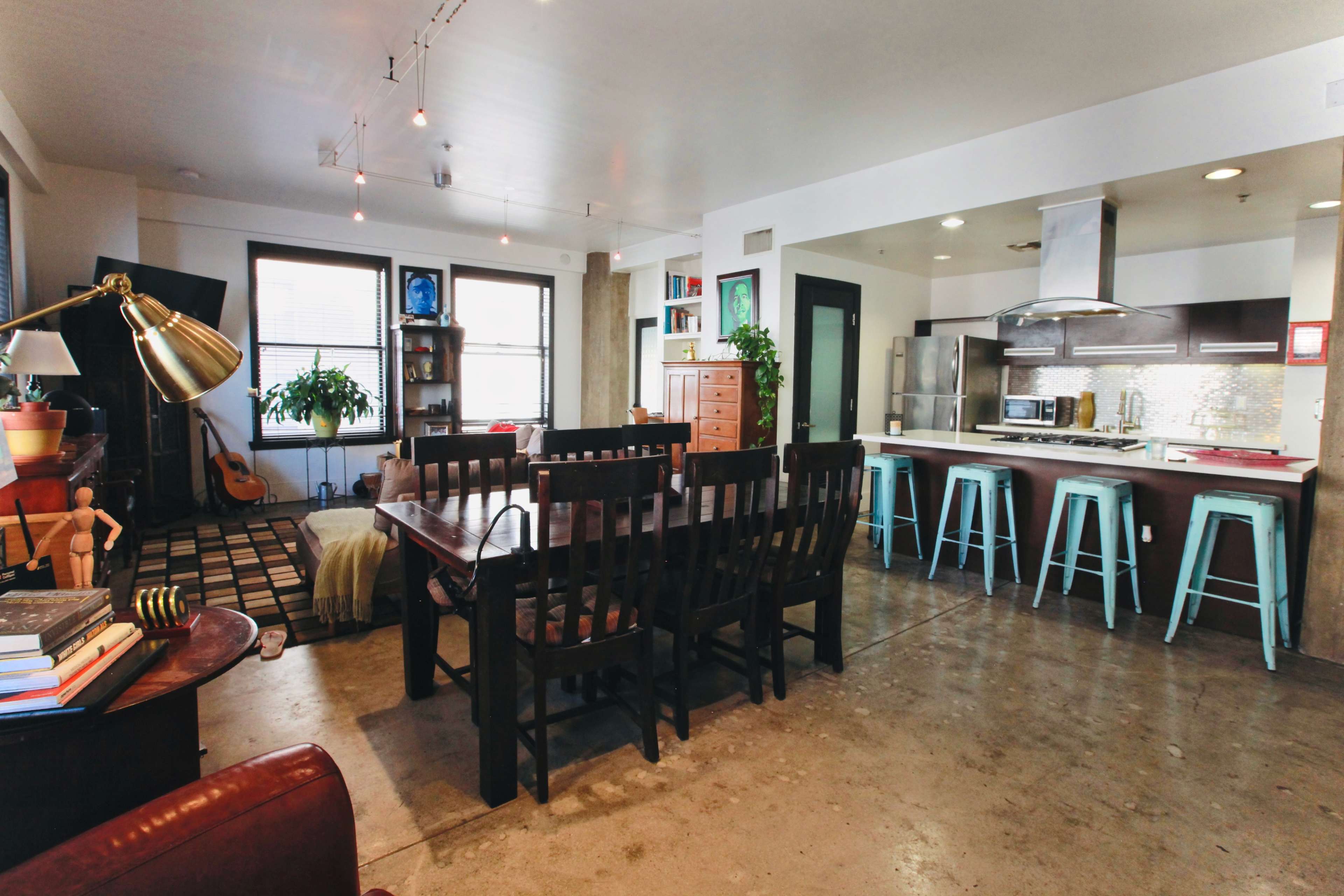 The image shows an open-concept living and dining area with a wooden dining table, kitchen with modern appliances, and a seating area featuring a guitar and plants.