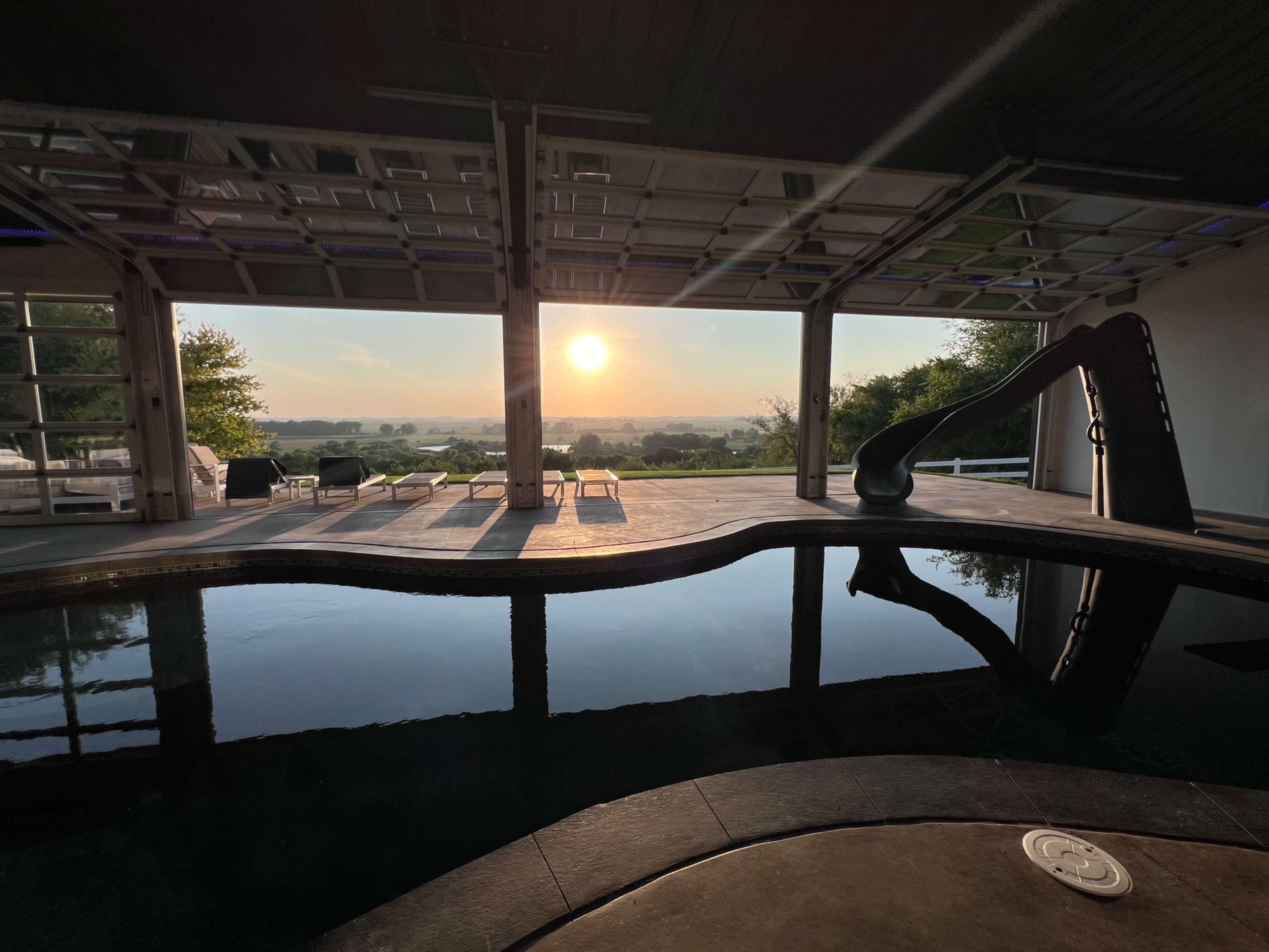 The image shows an indoor pool area with large windows that overlook a scenic landscape at sunset.