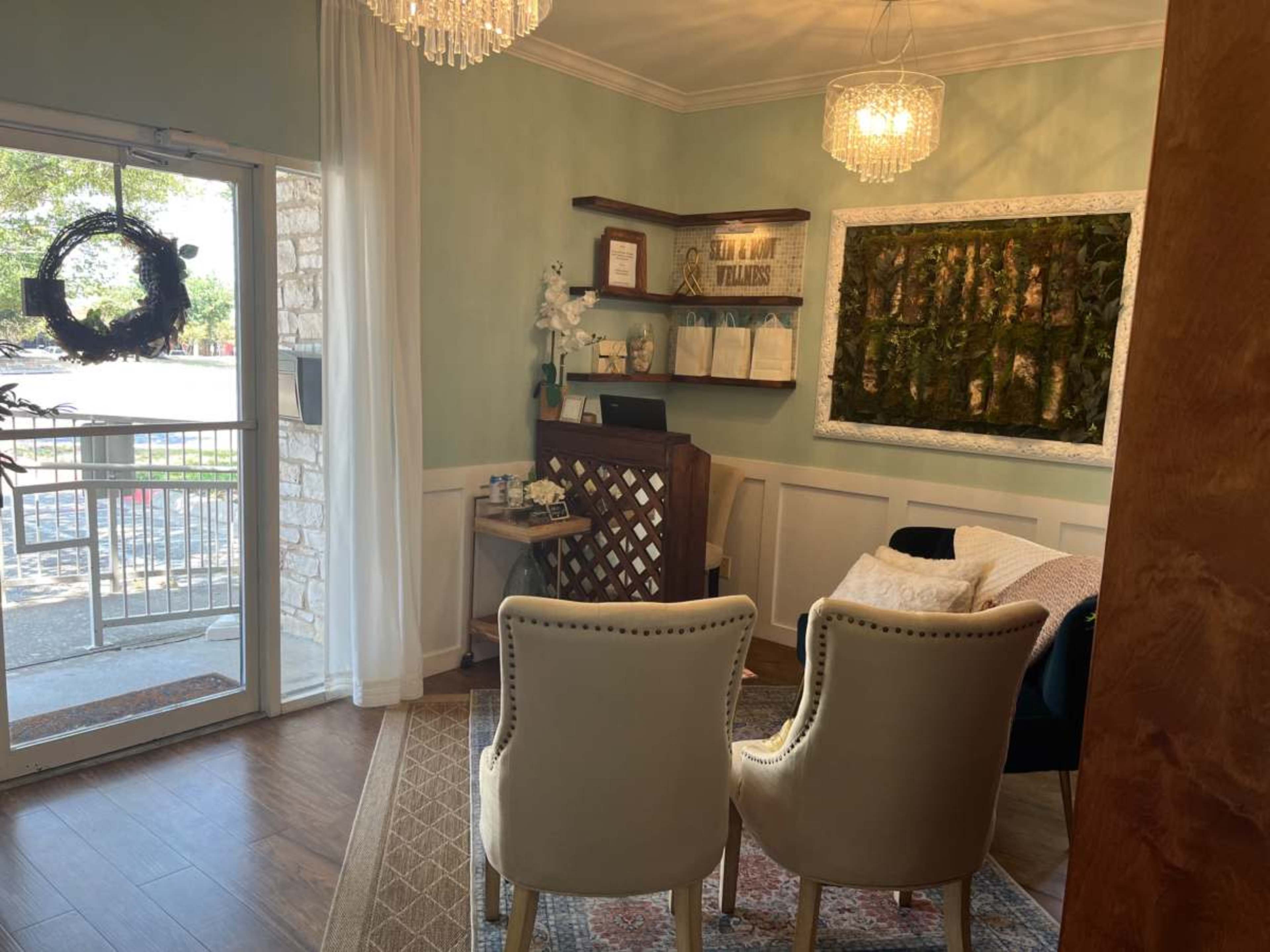 The image depicts a cozy waiting area with two upholstered chairs, a wooden reception desk, and a view of an entrance with a glass door.