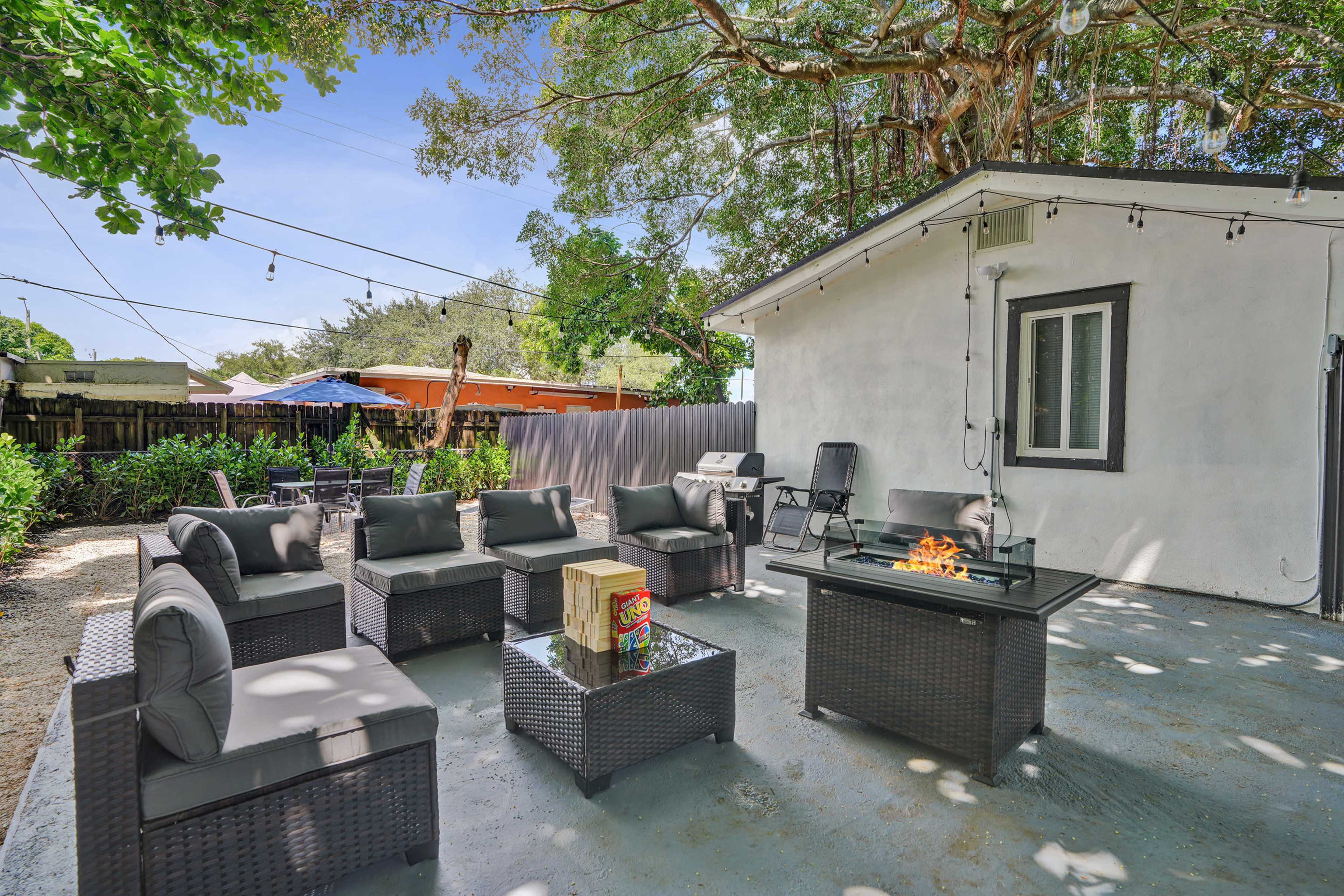 The image shows a cozy outdoor patio area with a seating arrangement around a fire pit, surrounded by greenery and a small building in the background.