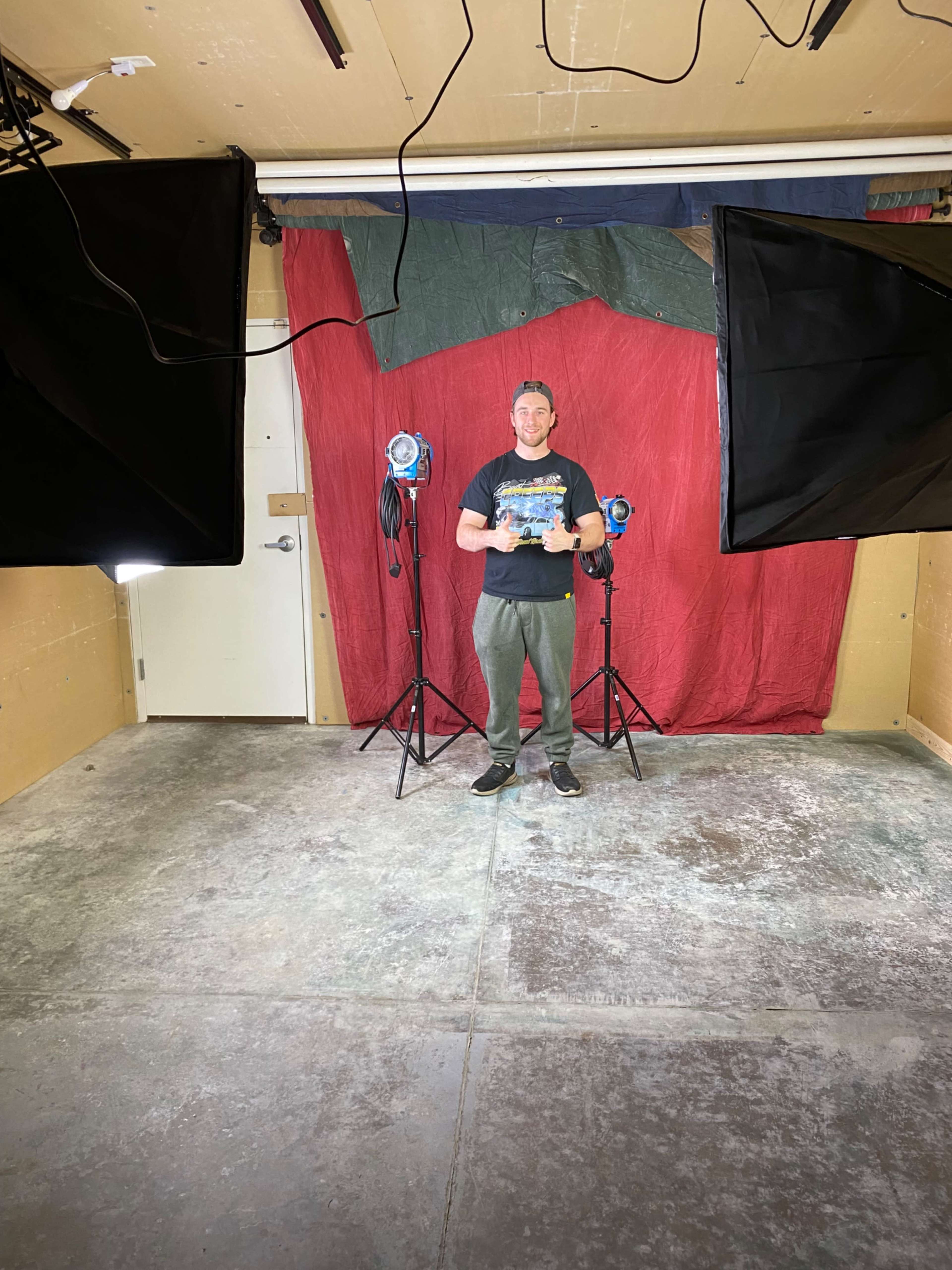 A person stands in a studio space with two lighting setups and a red backdrop.