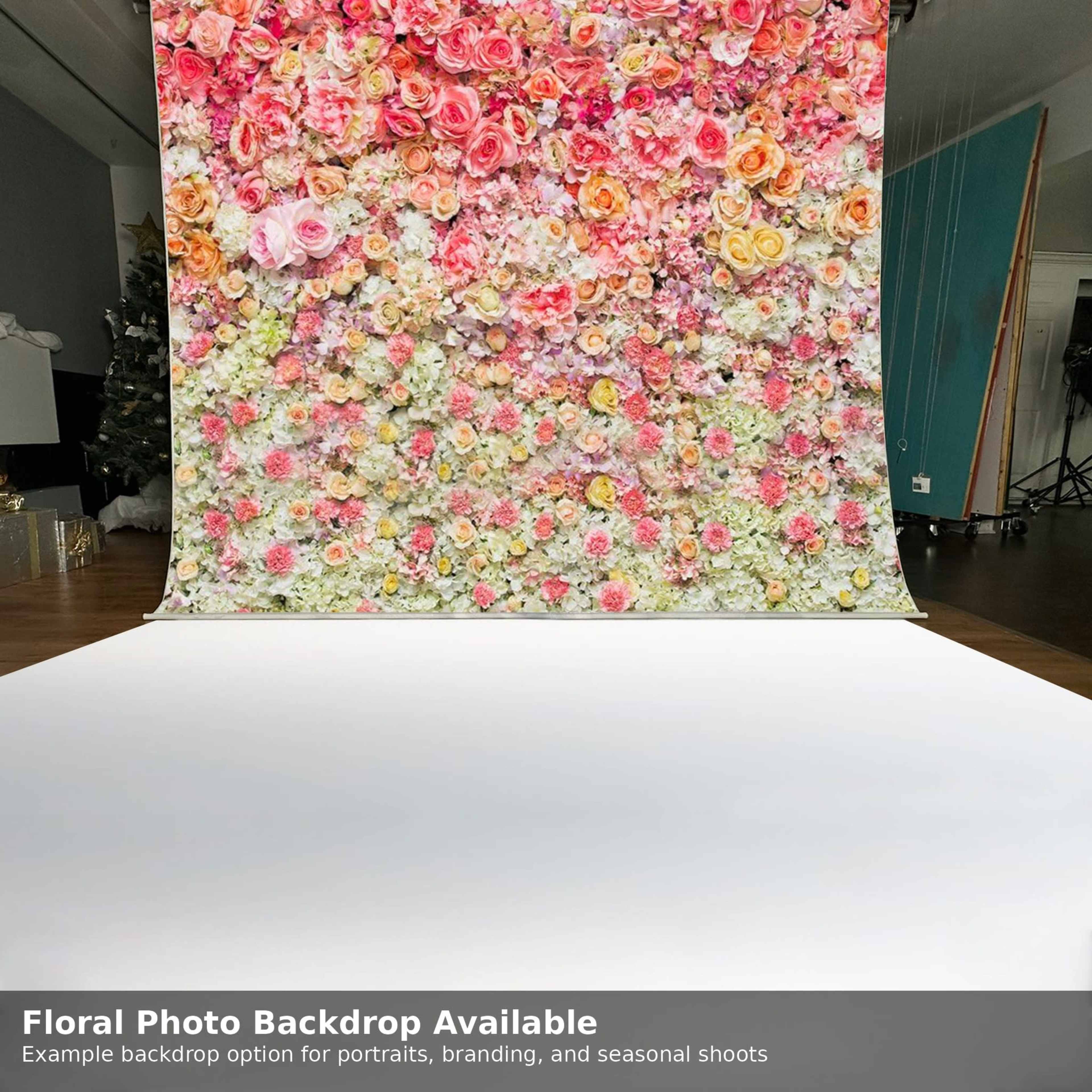 The image features a large floral backdrop filled with colorful roses and other flowers, set up in a photography studio with a blank white floor.