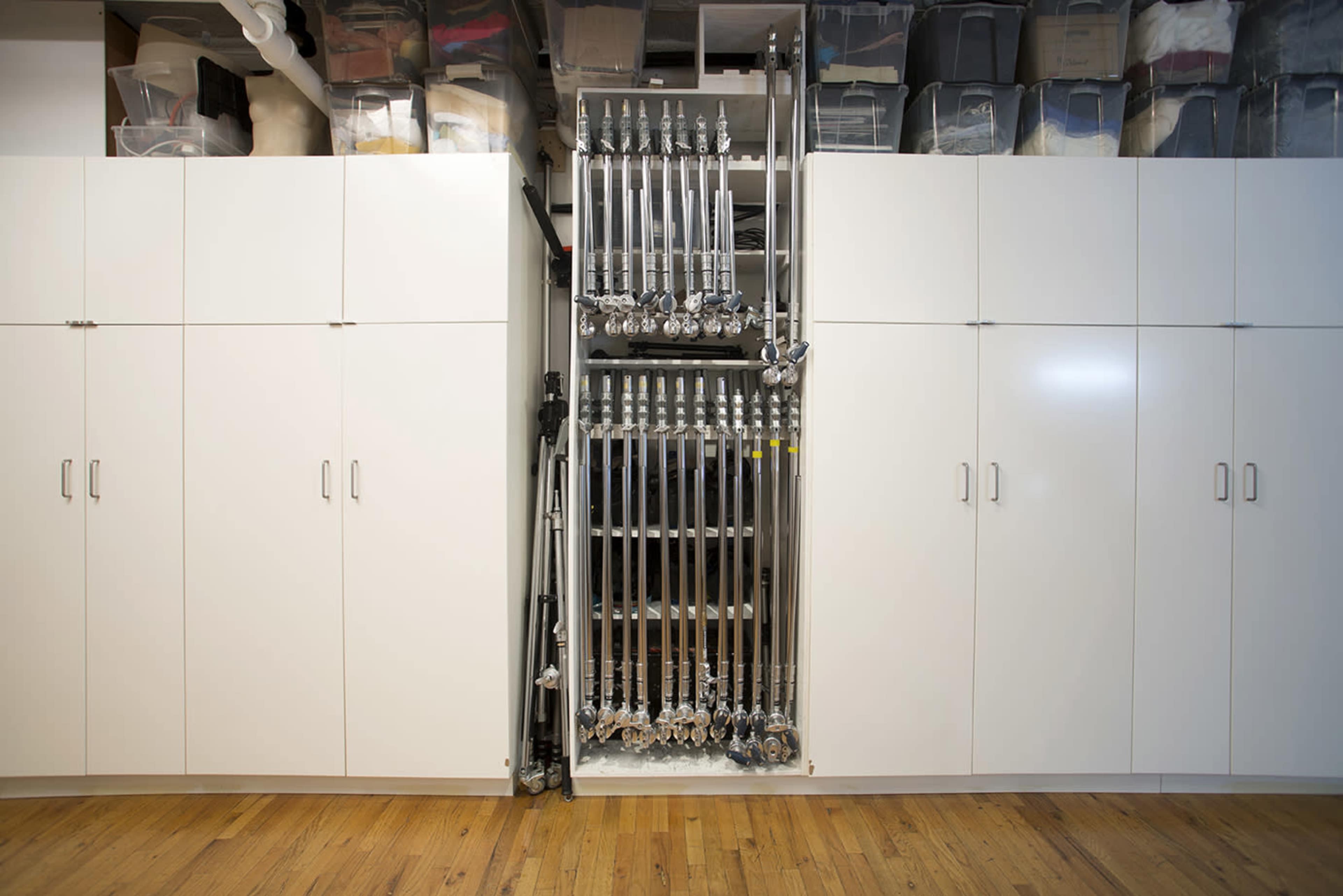 The image shows a neatly organized storage area with white cabinets flanking a metal rack holding silver equipment, surrounded by plastic bins in the upper section.
