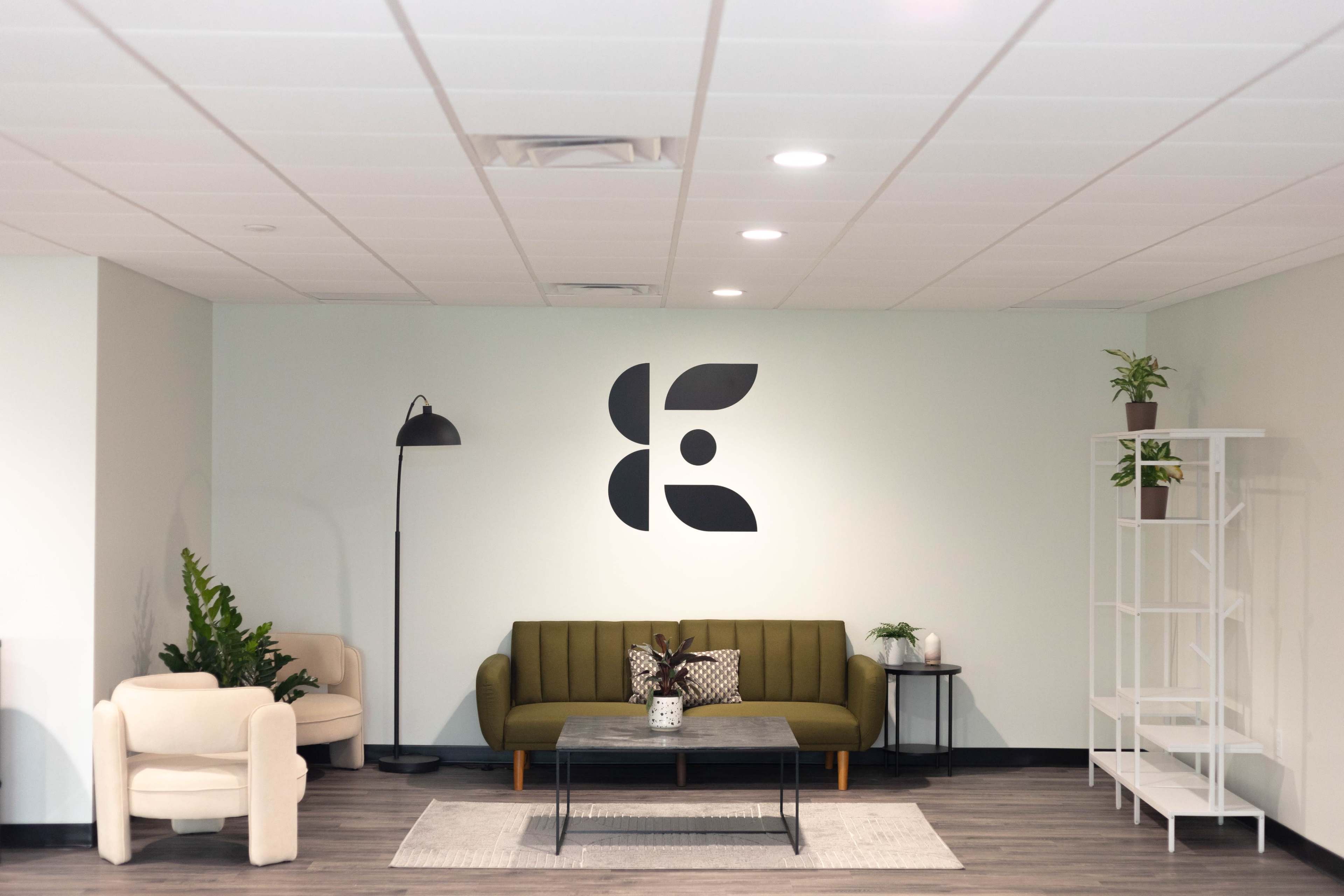 A modern waiting area features a green couch, a white armchair, a coffee table, and a shelf with plants against a light blue wall displaying a large black logo.