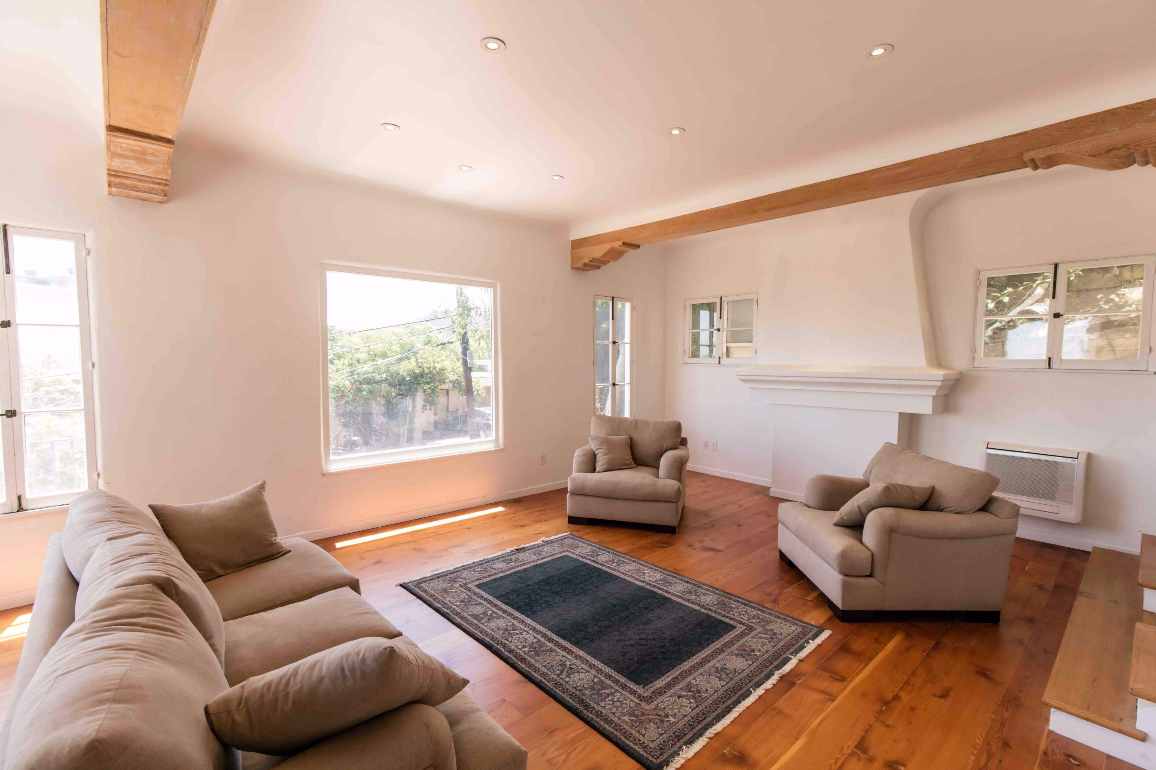 A bright, minimalist living room features two armchairs and a sofa arranged around a central rug, with large windows allowing natural light to enter.