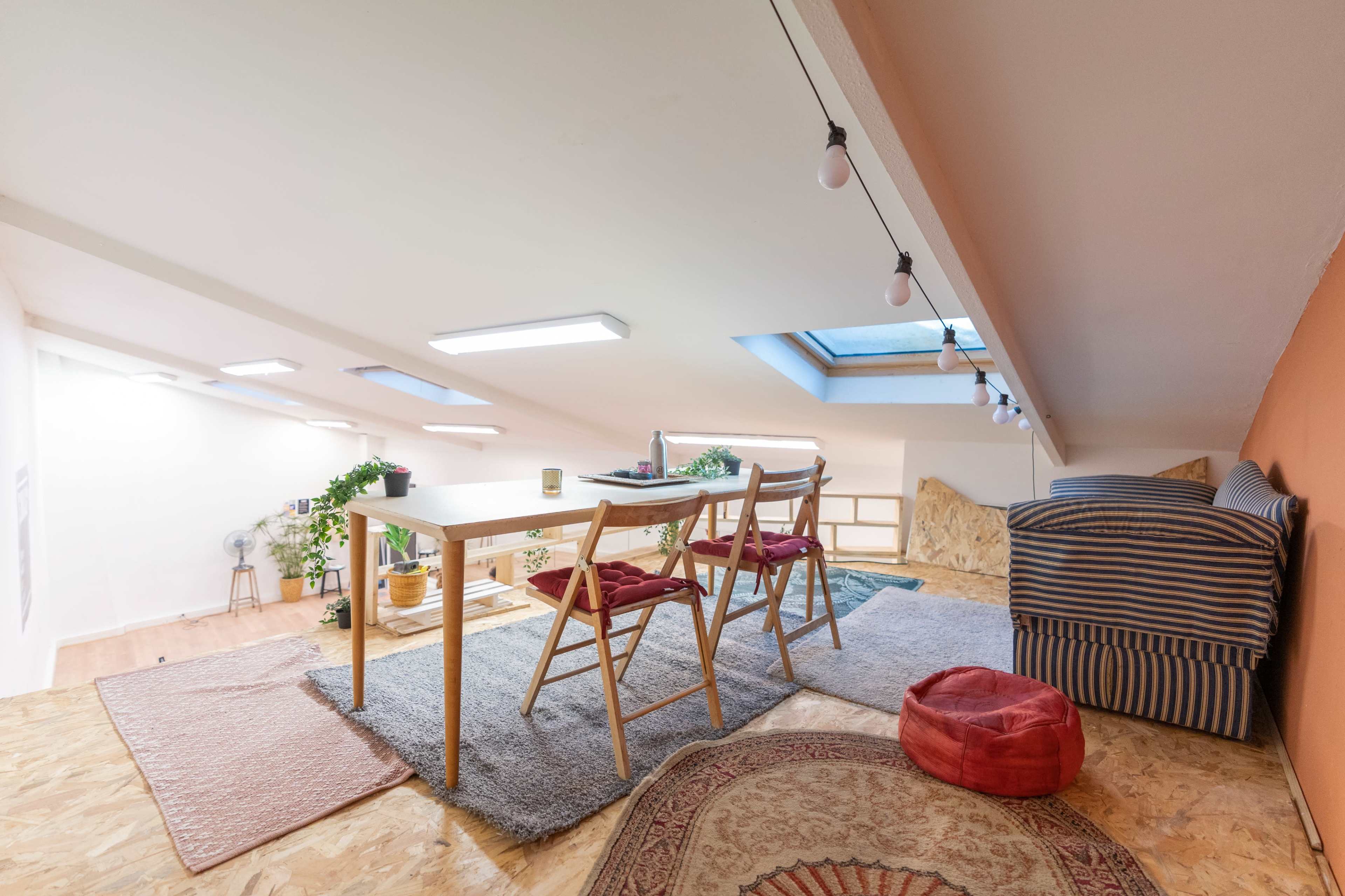 A cozy loft space featuring a wooden table with four chairs, a striped sofa, and potted plants under a skylight.