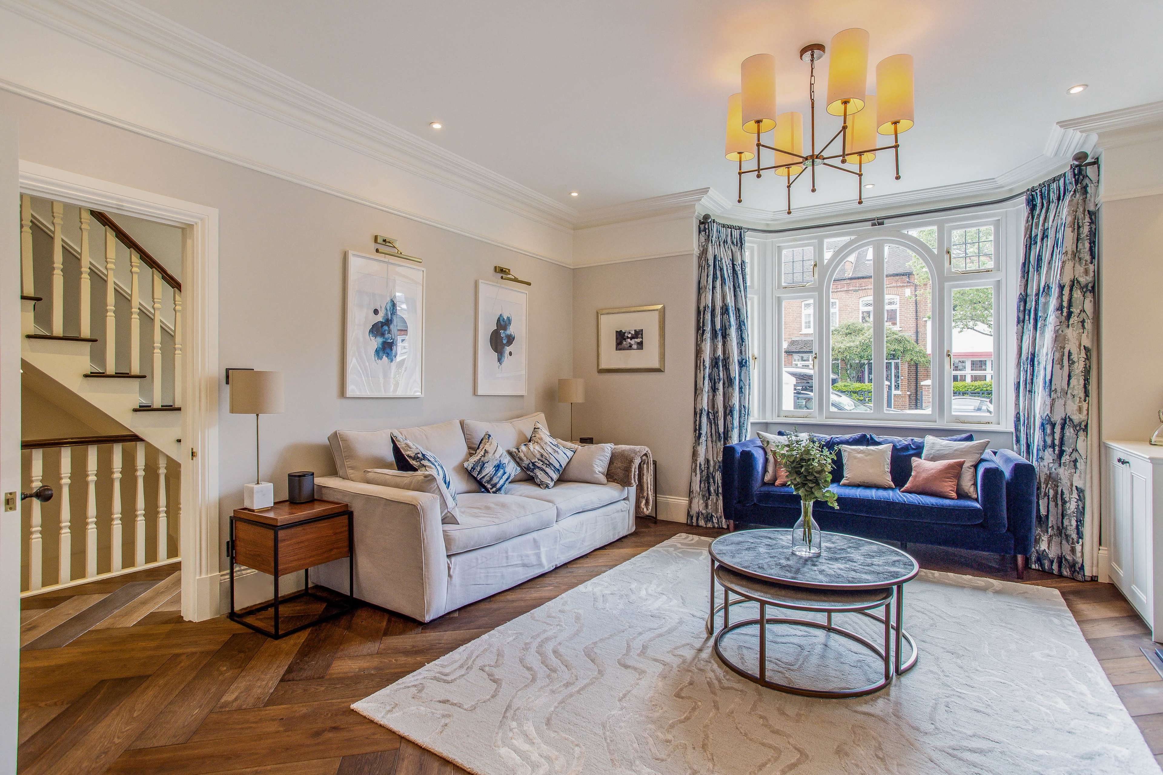 A well-furnished living room features a light-colored sofa, a blue armchair, a round coffee table, and large windows dressed with patterned drapes.