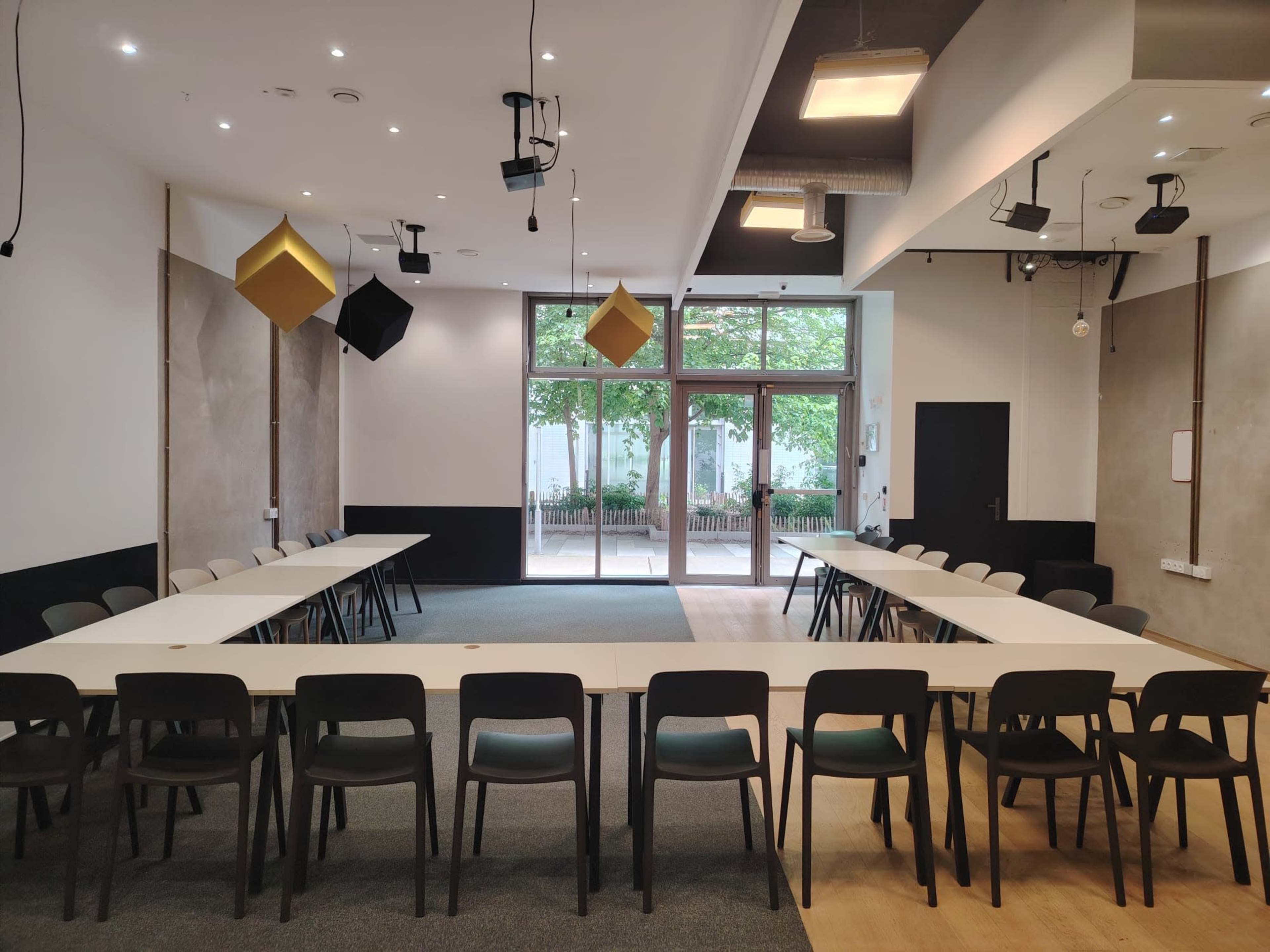 A conference room features a large U-shaped table surrounded by black chairs, with large windows providing a view of outdoor greenery.