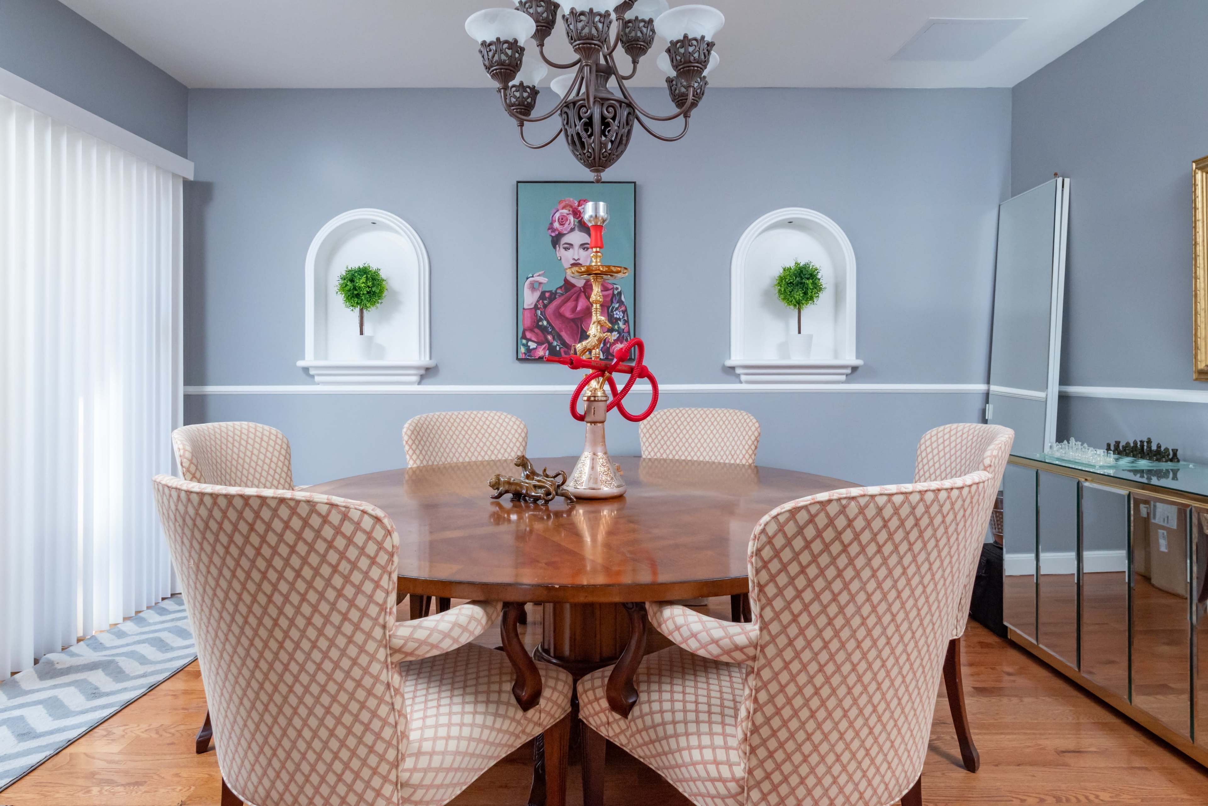 A round wooden dining table surrounded by upholstered chairs is set in a modern dining room featuring wall art and decorative alcoves.