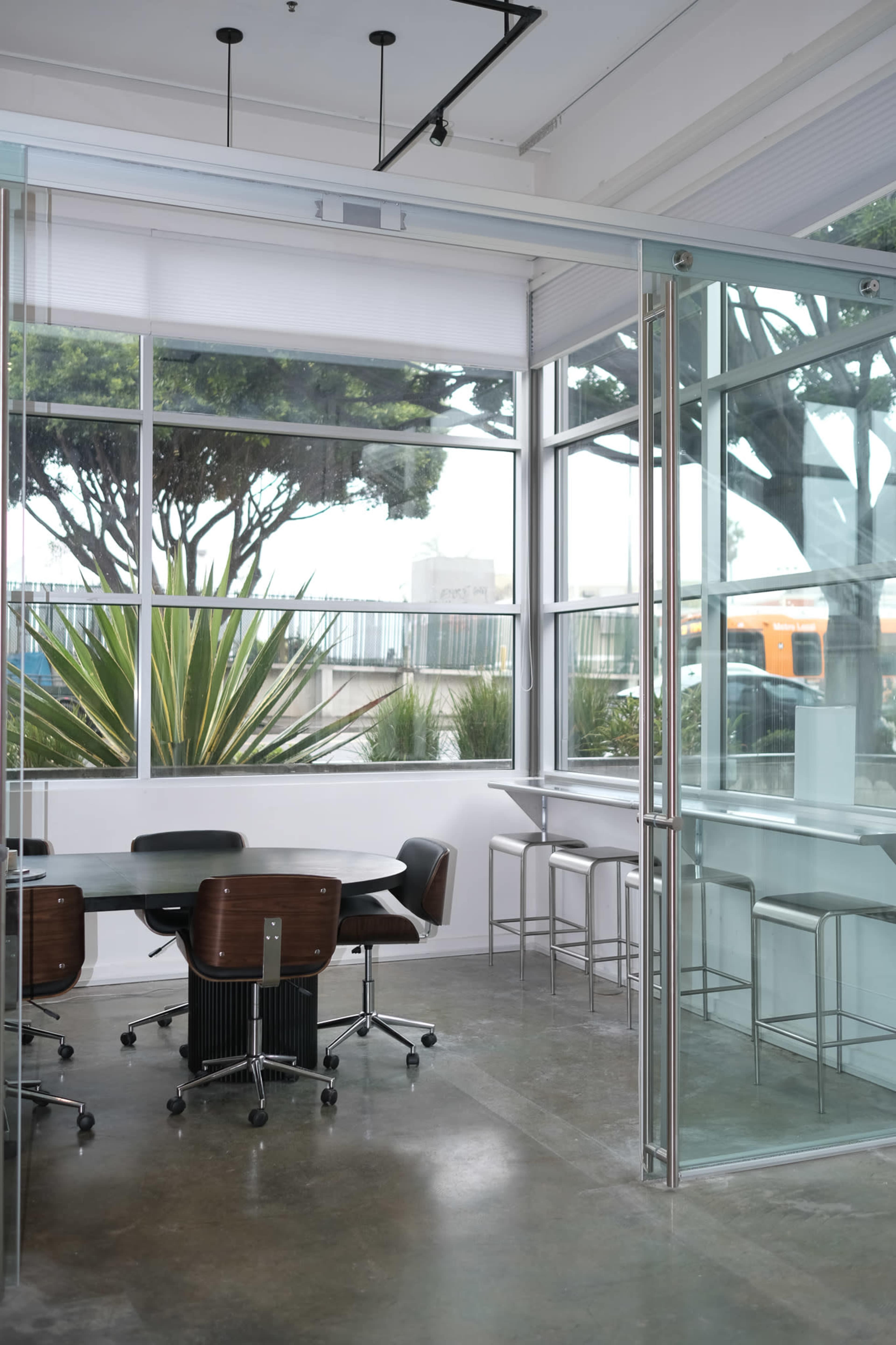 A modern office space features a glass-walled meeting room with a round table and chairs, alongside a bar-height seating area near large windows.