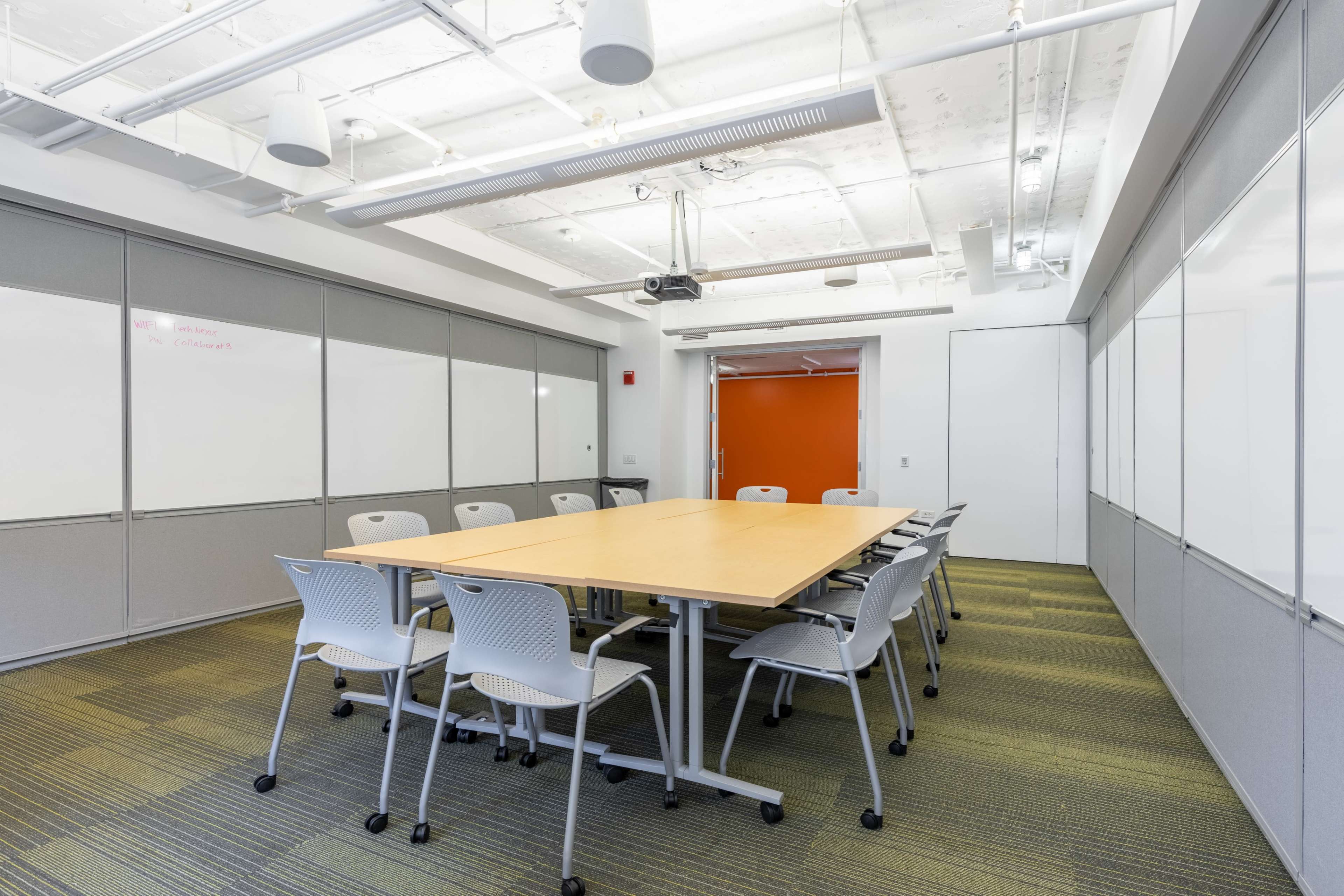 A conference room features a large table surrounded by office chairs, with whiteboards on the walls and an orange door at the back.