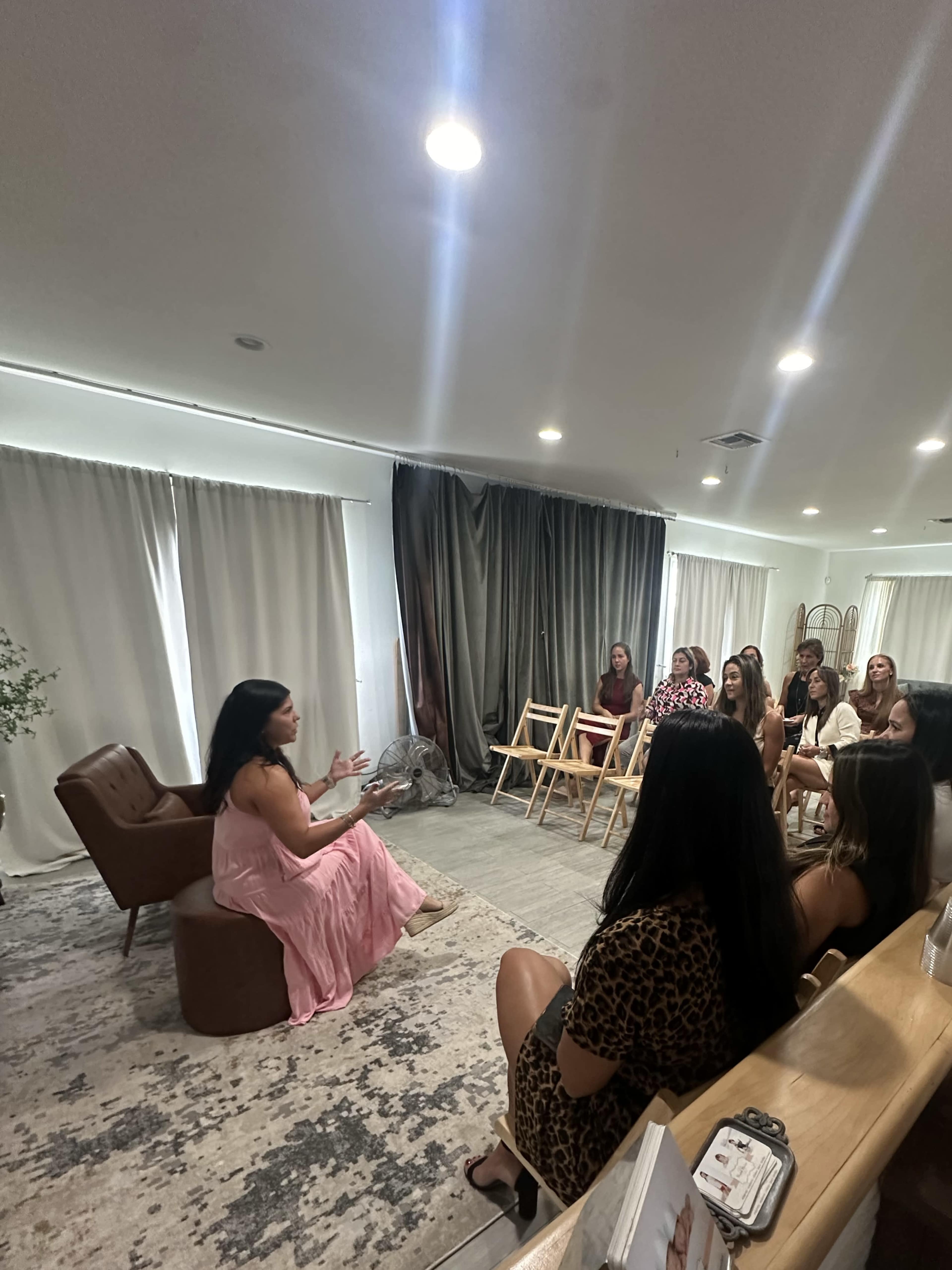 A woman in a pink dress speaks to a group of seated individuals in a softly lit room with neutral-colored curtains and a rug.
