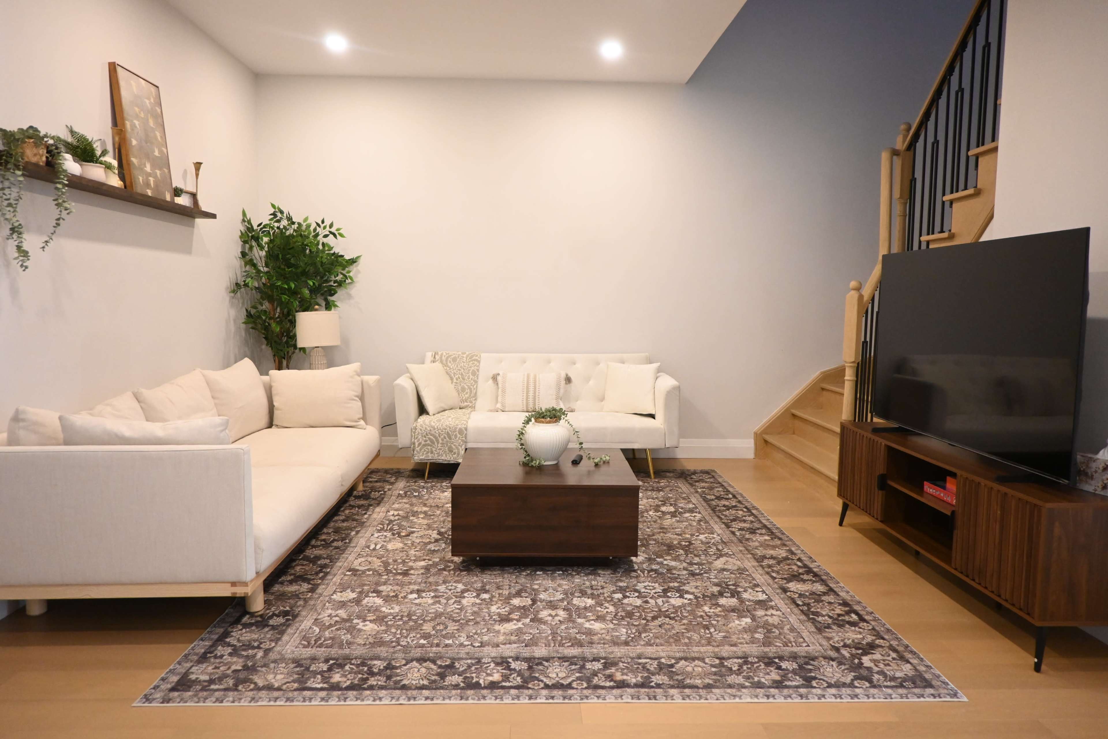 The image shows a modern living room featuring a white sectional sofa, a light-colored armchair, a wooden coffee table on a patterned rug, and a television on a low wooden console, with a staircase in the background and decorative plants.
