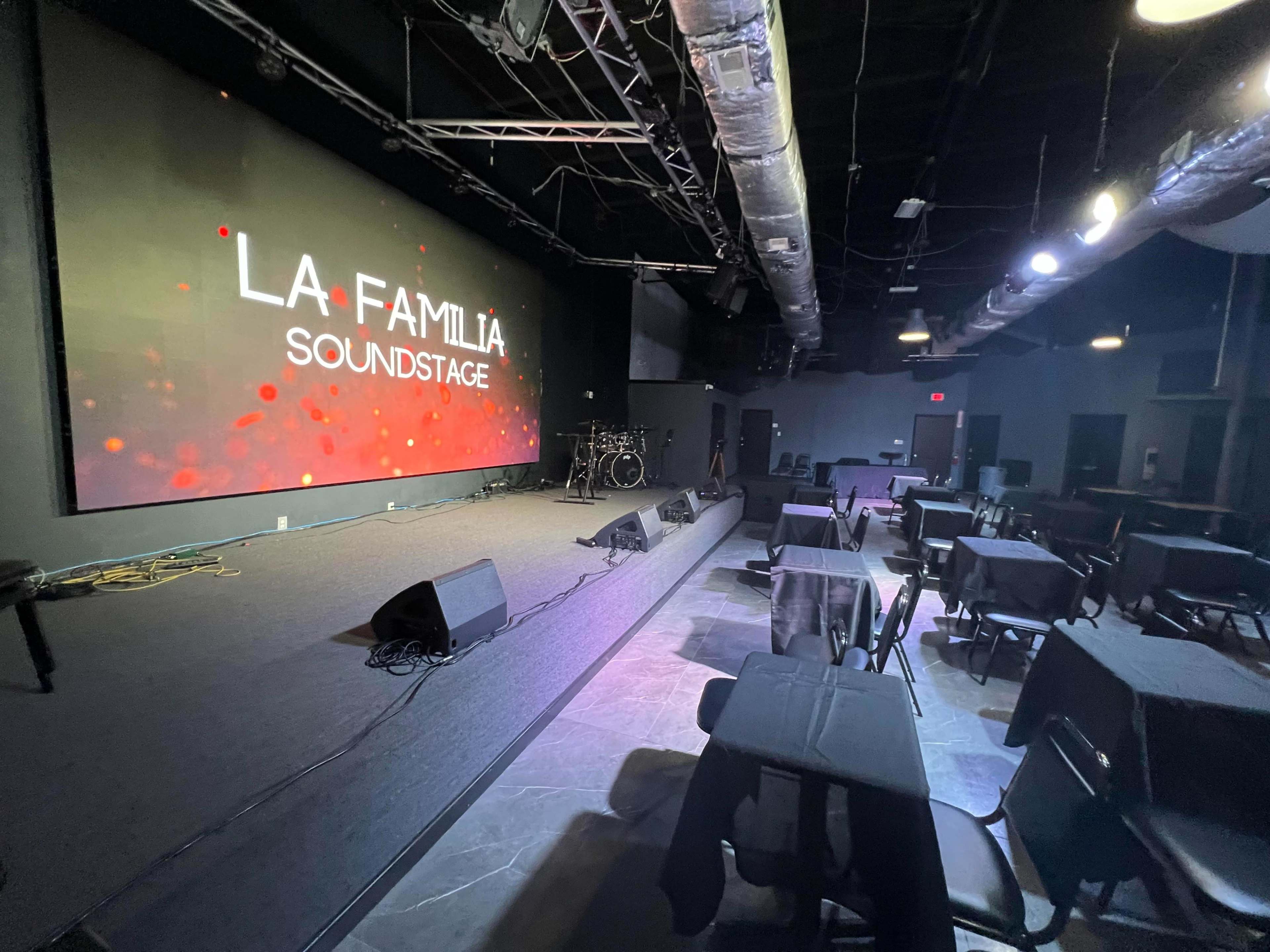 A spacious performance venue features a large screen displaying "LA FAMILIA SOUNDSTAGE," with tables set for an audience in front of a stage and music setup.