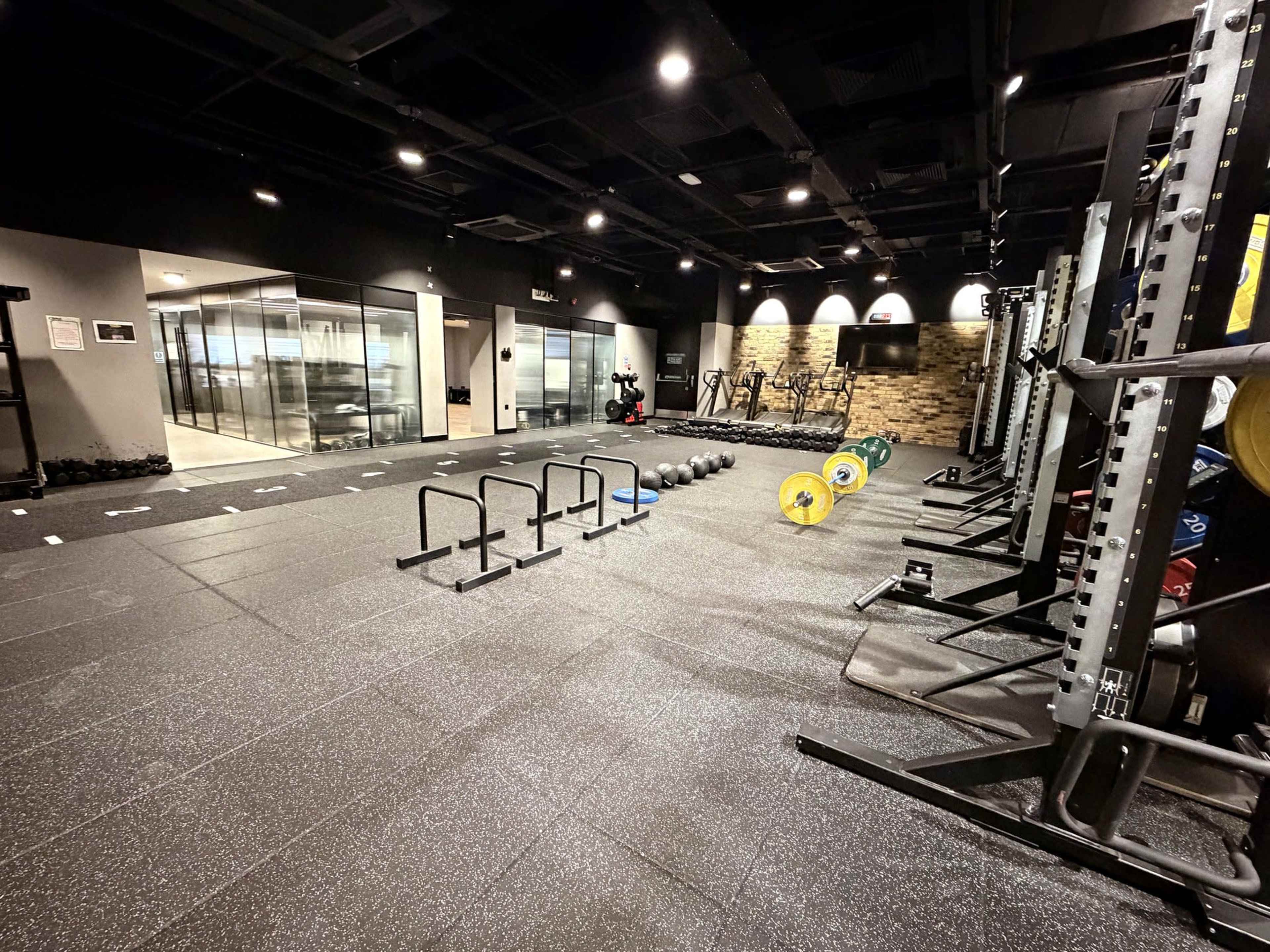 The image shows a modern gym interior with weightlifting equipment, kettlebells, and a designated workout area on a rubber floor.