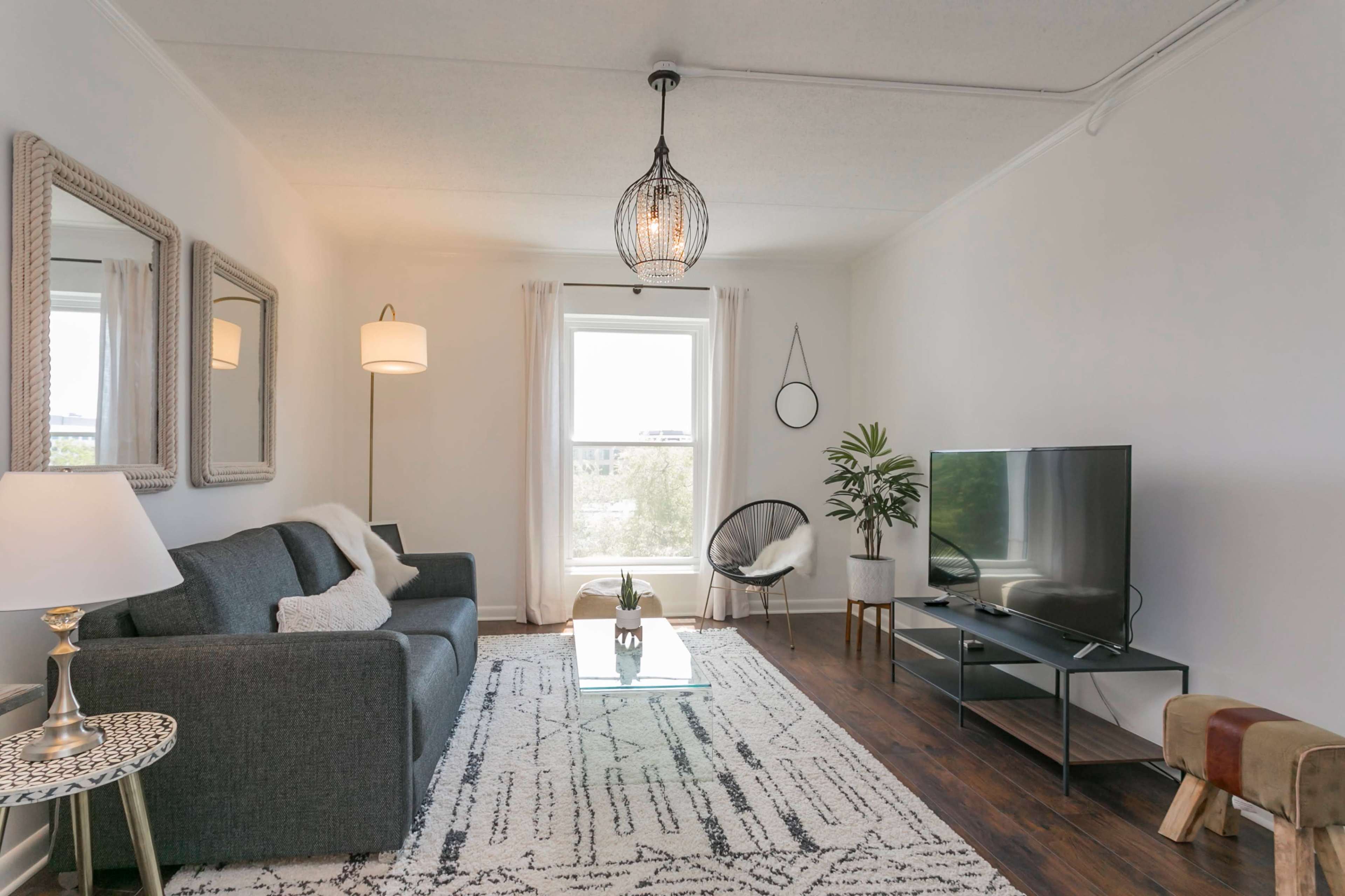 A modern living room features a gray sofa, a glass coffee table, a TV on a black stand, and a large window with natural light.