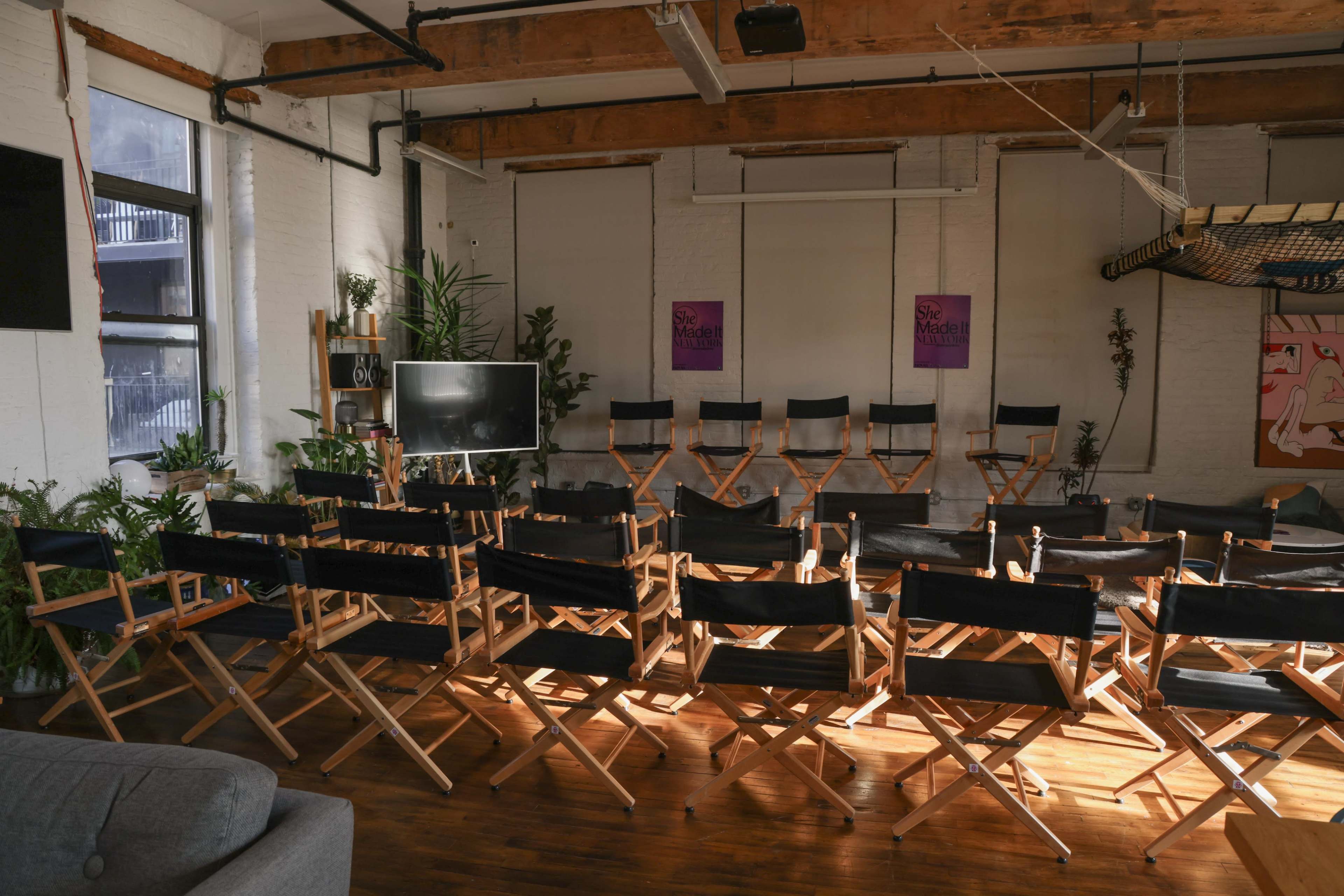 A room with wooden beams featuring several rows of empty director's chairs facing a television and adorned with potted plants.
