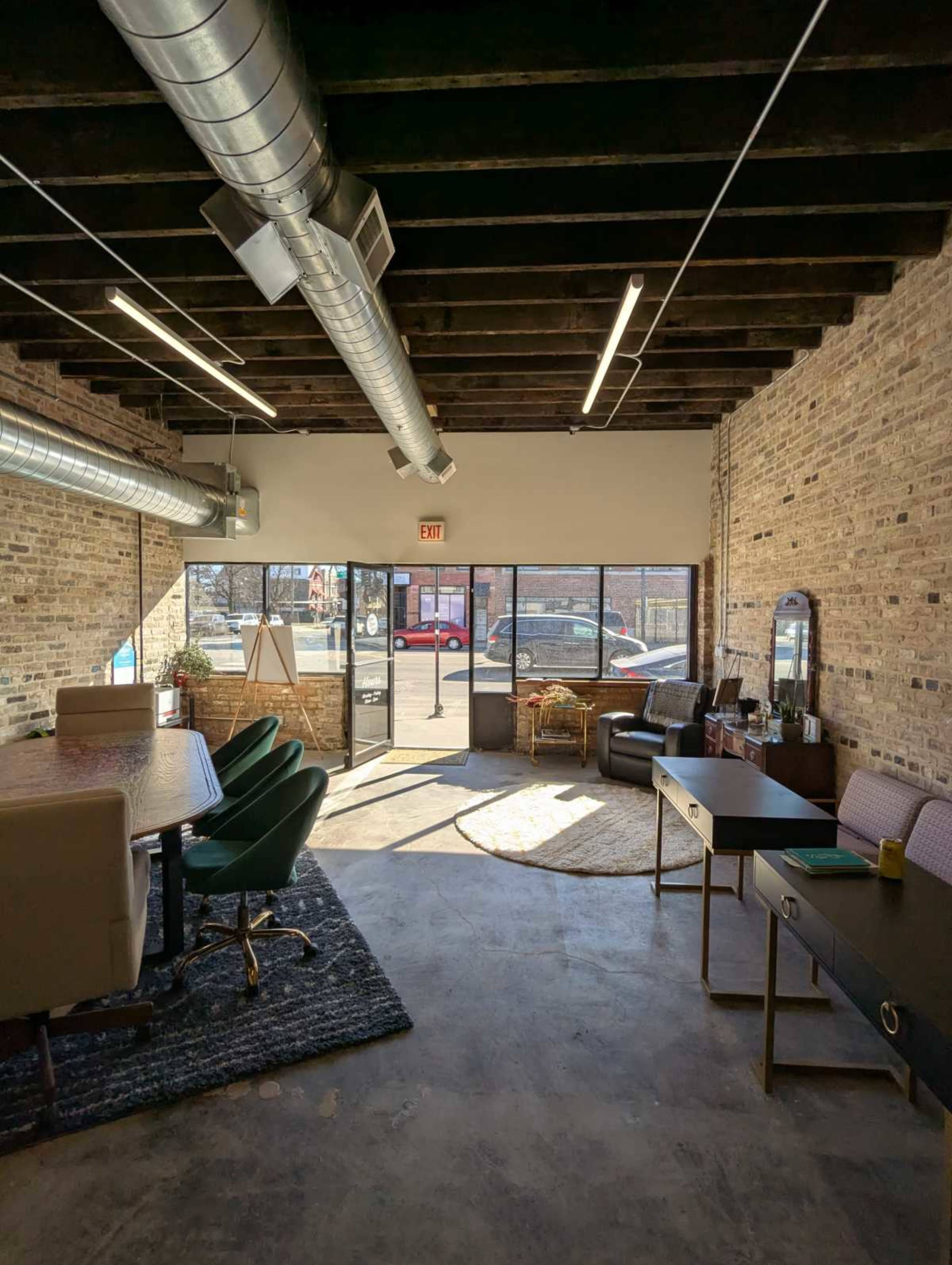The image shows a spacious, modern office interior with exposed brick walls, large windows letting in natural light, and a mix of seating and workspace areas.